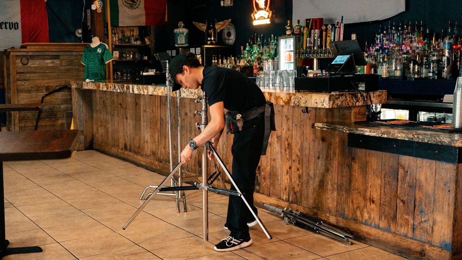 A bartender mops the floor of an empty bar with wooden paneling and liquor bottles on shelves.
