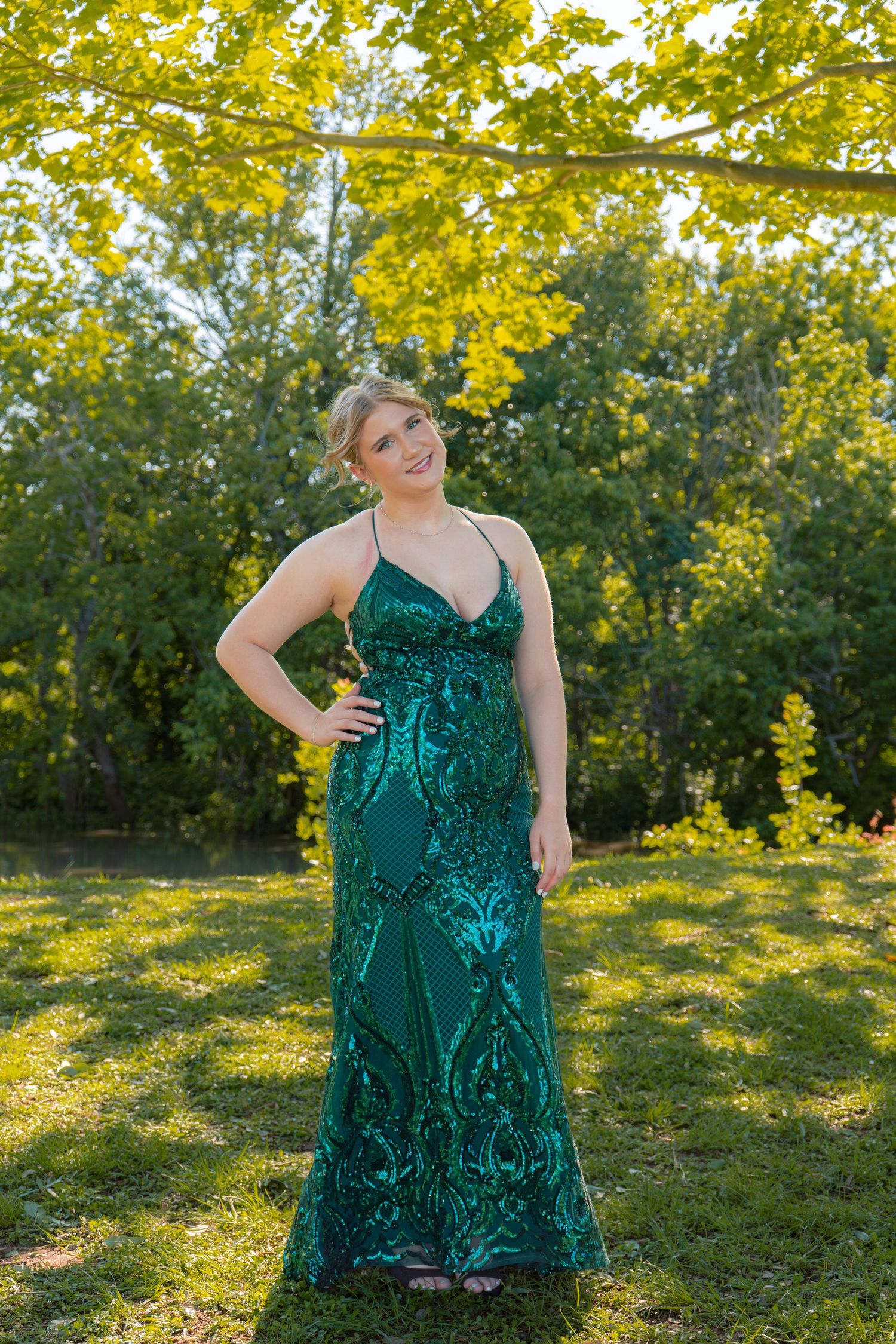 Woman in stunning emerald green sequin formal gown poses outdoors among green trees at sunset.