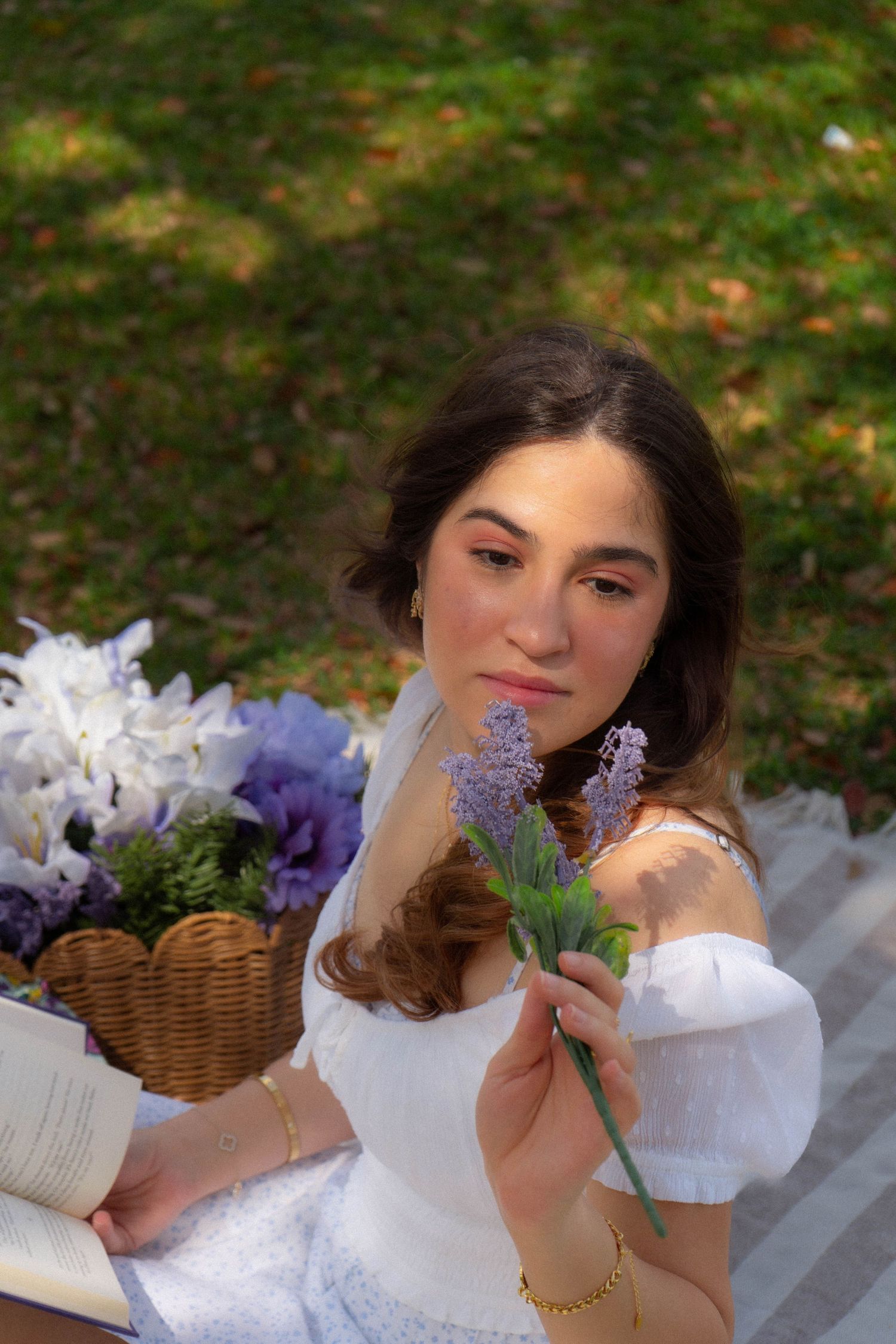 Young woman in white off-shoulder dress holding lavender flowers at park picnic, looking at camera.