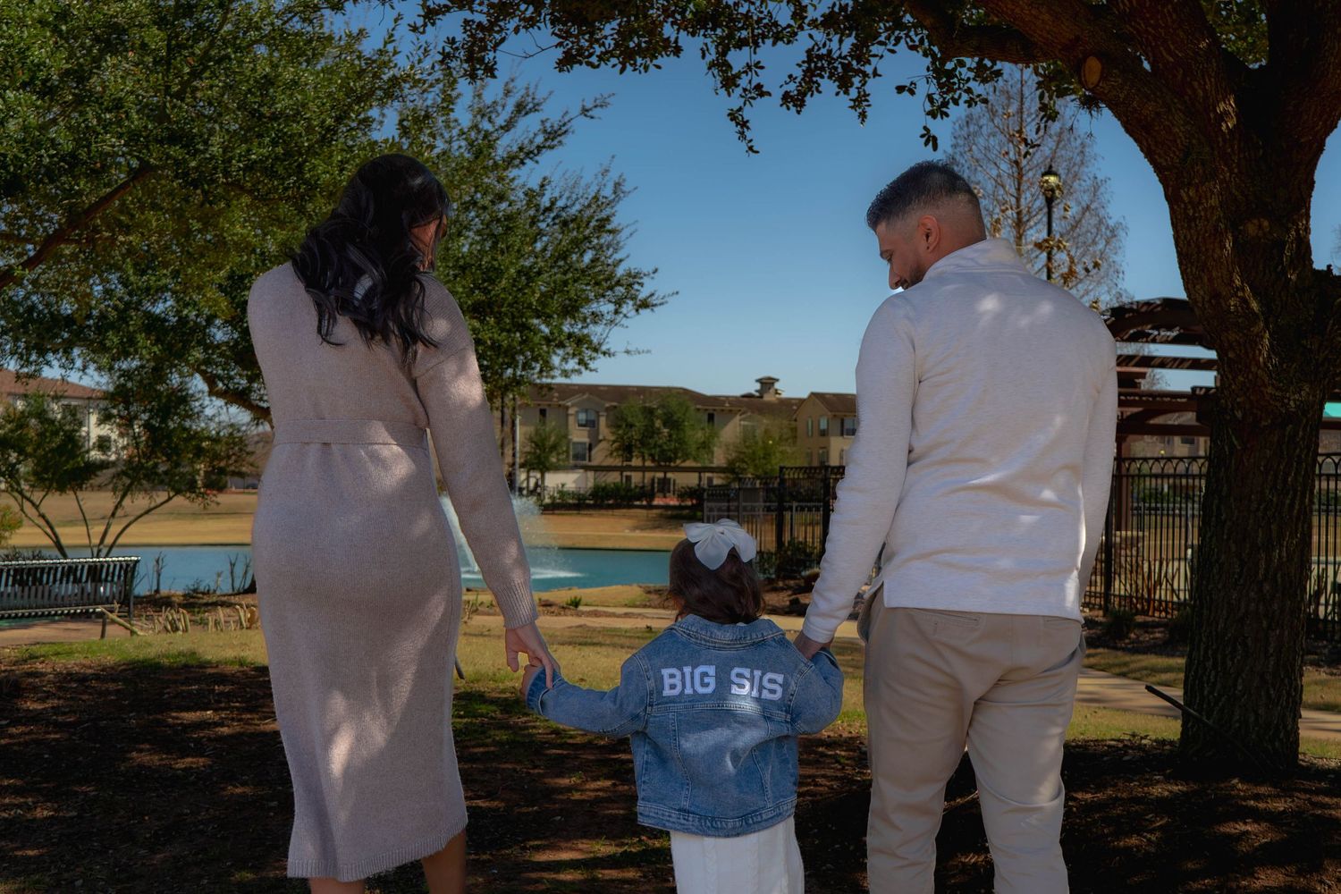 Family maternity photo from behind showing toddler in Big Sis denim jacket holding parents hands.
