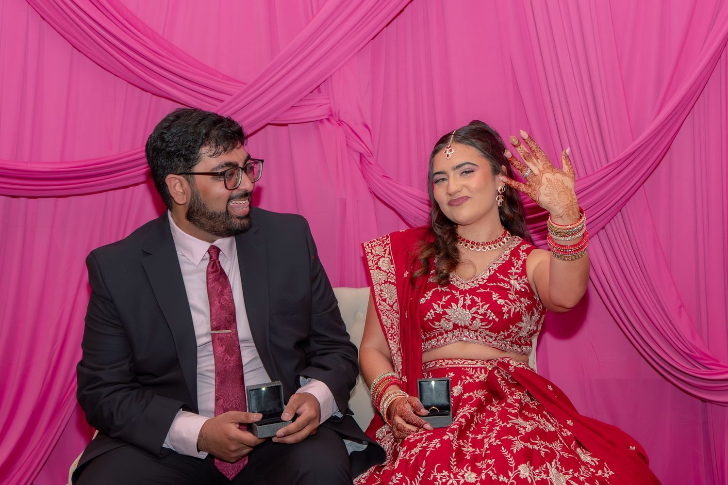 South Asian couple seated before pink backdrop, bride in red lehenga waving while groom in black suit holds ring box.