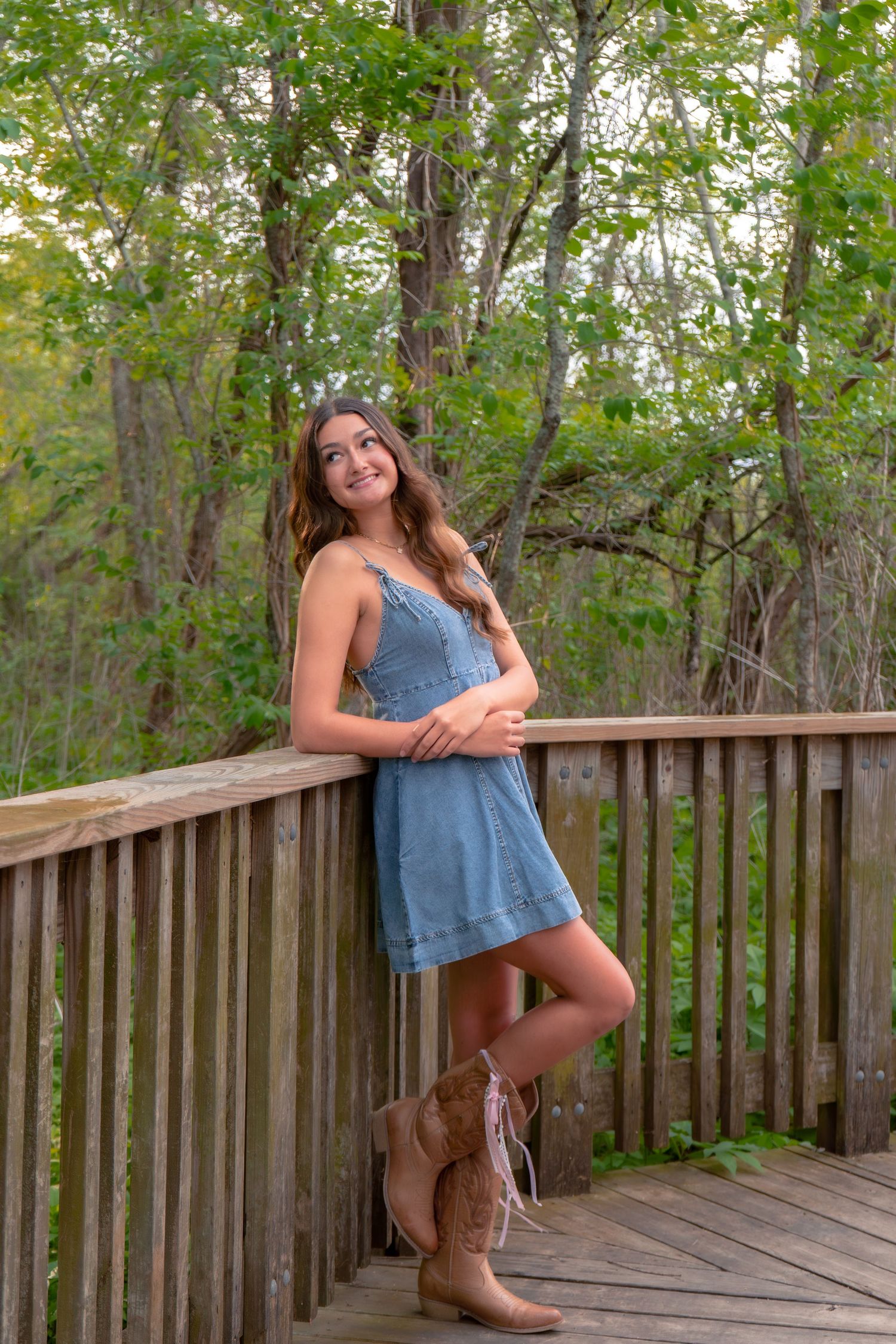 Young woman in denim dress and cowboy boots smiling on wooden deck surrounded by lush green trees.