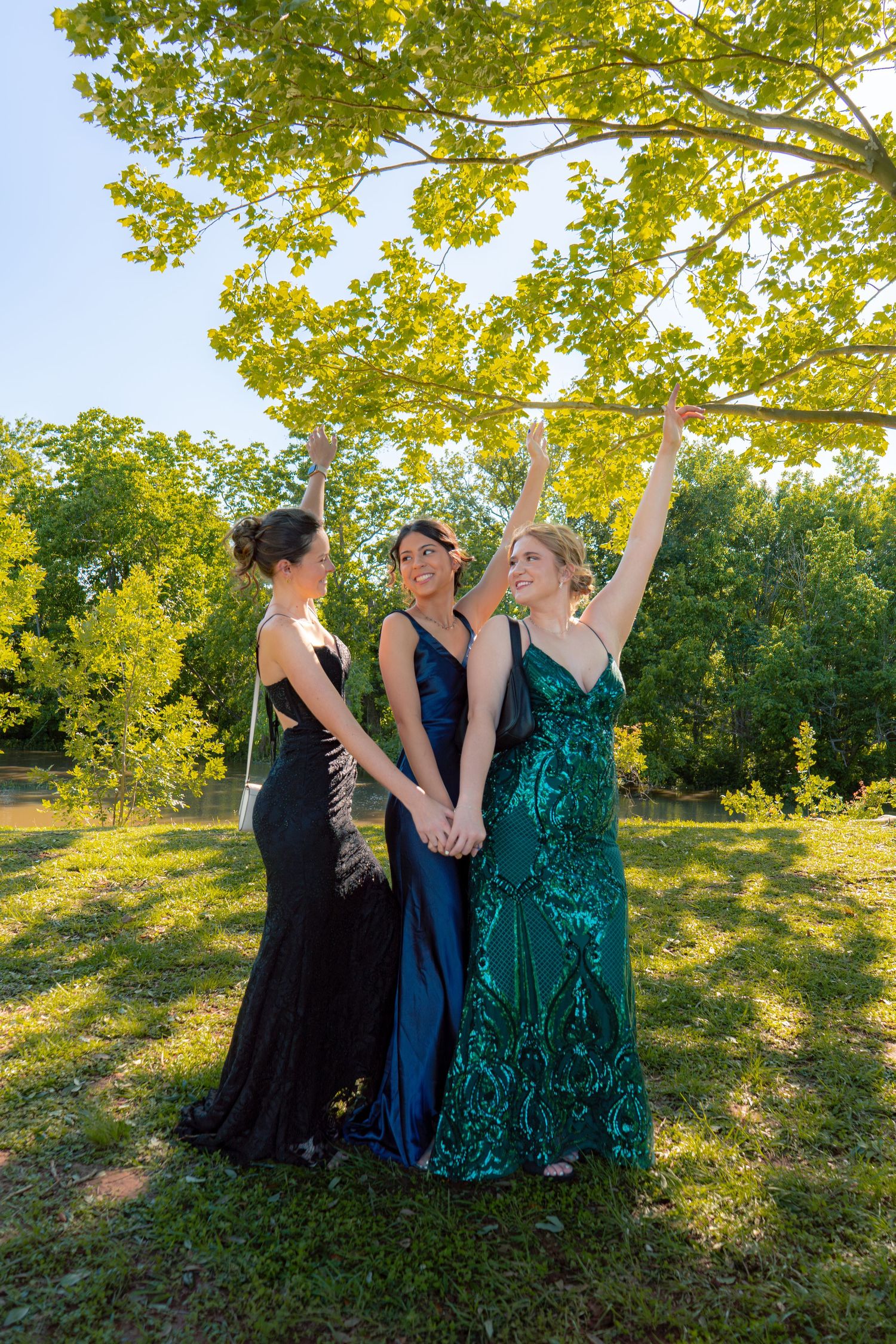 Three young women in elegant formal gowns laugh and raise their arms while holding hands outdoors under a tree.