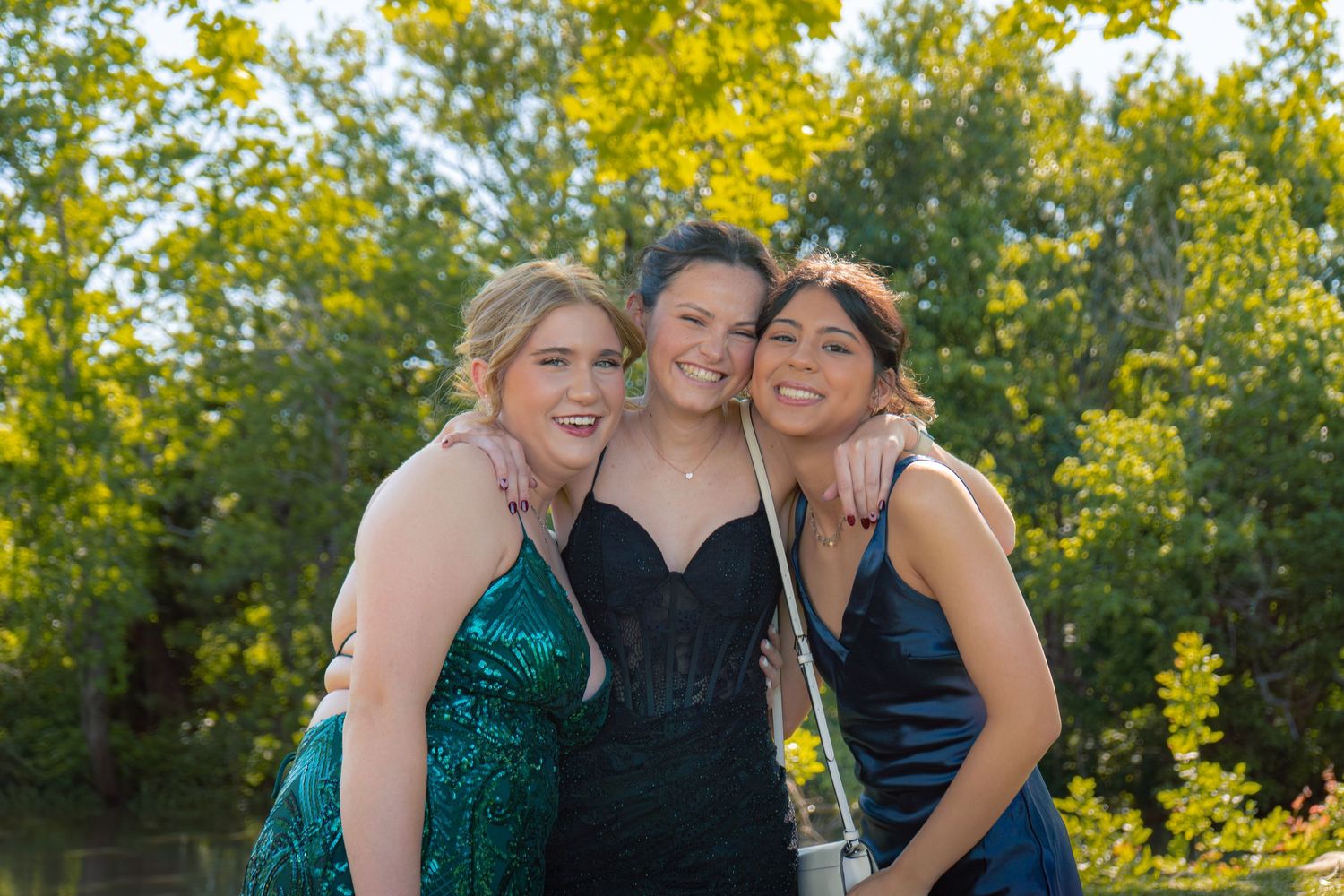 Three teenage girls in elegant prom dresses pose together outdoors with lush green trees in the background.