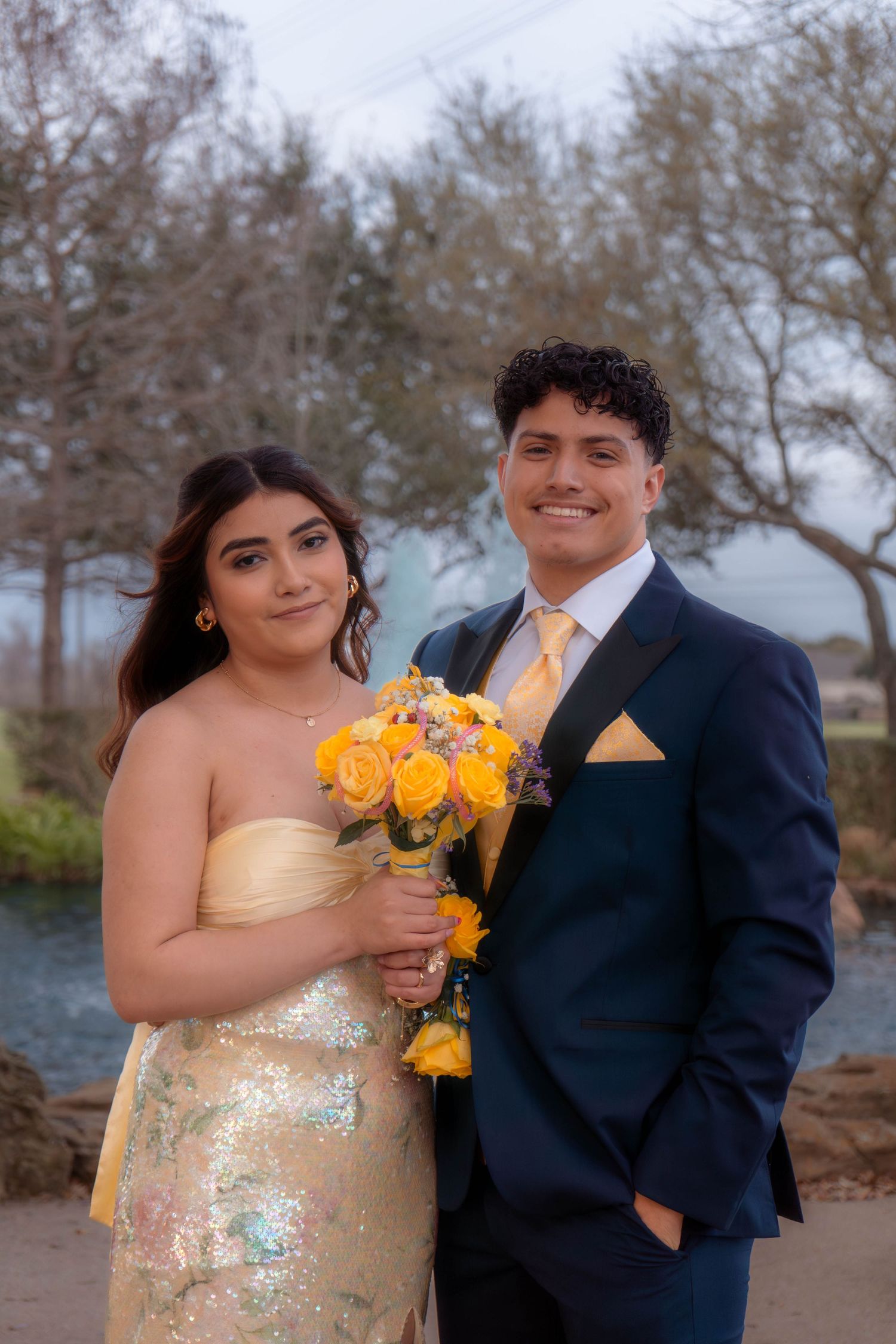 Prom couple posing outdoors in formal wear with yellow rose bouquet and corsage.