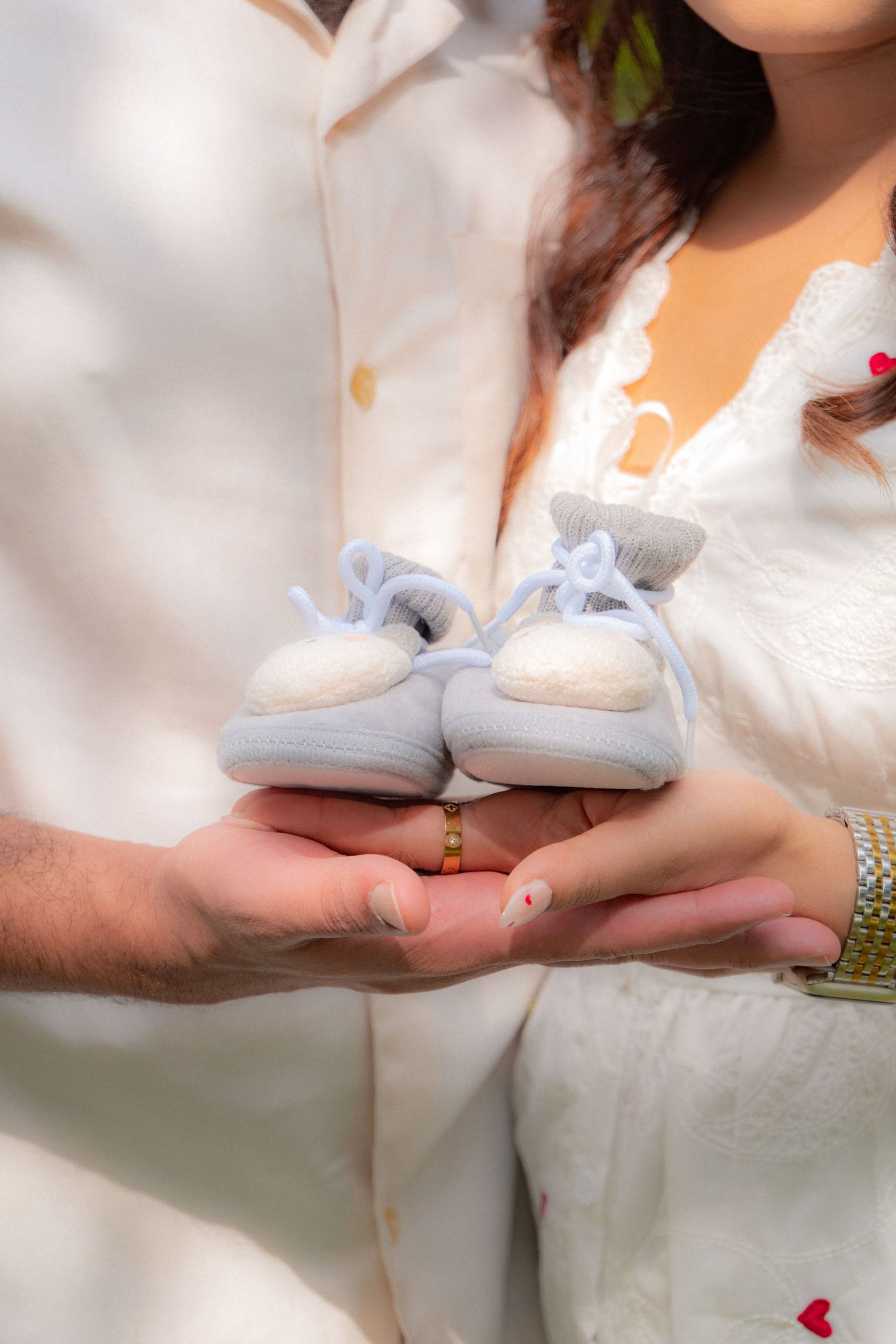 Expecting couple holds tiny blue baby booties together in their hands outdoors in soft sunlight.