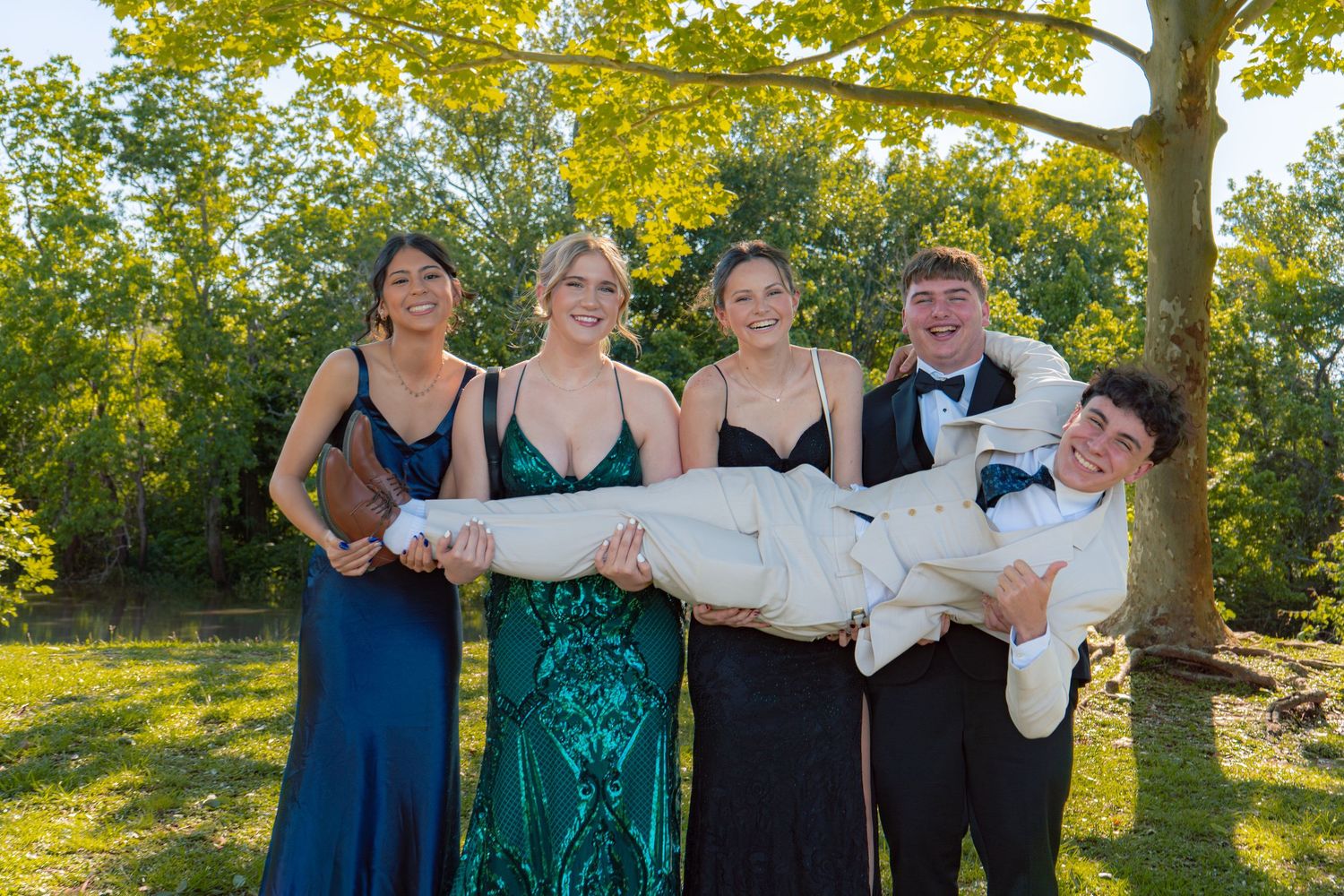 Four teens in formal wear carry a friend outdoors at prom, smiling in front of lush green trees.