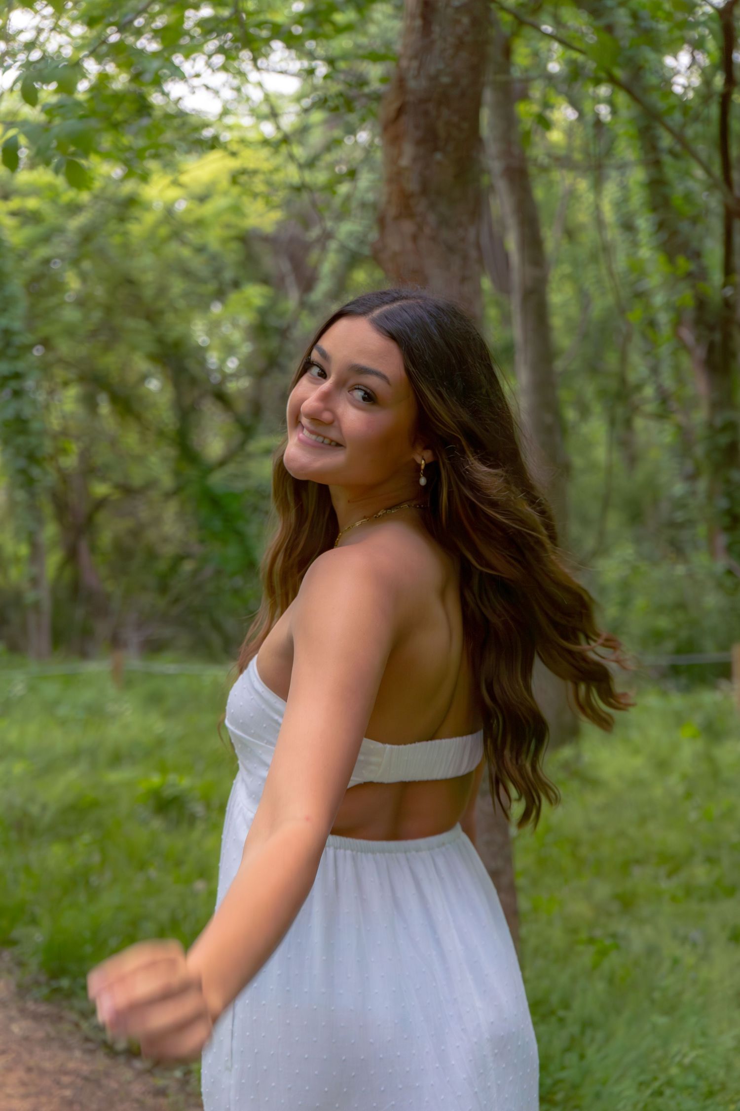 Young woman in white backless dress smiling over shoulder in lush green forest setting.