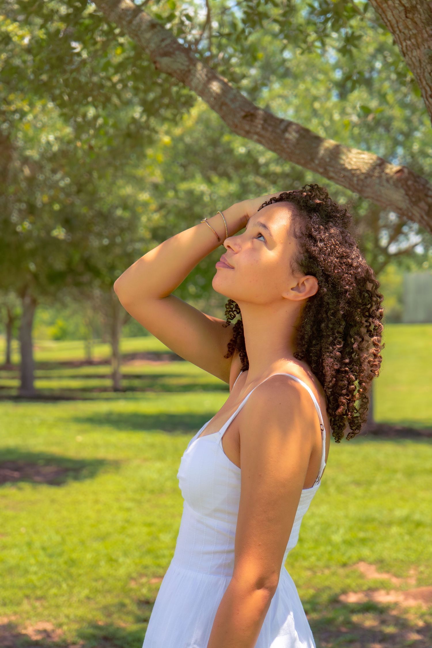 Young woman with curly hair in white dress looks up gracefully in a sunny green park.