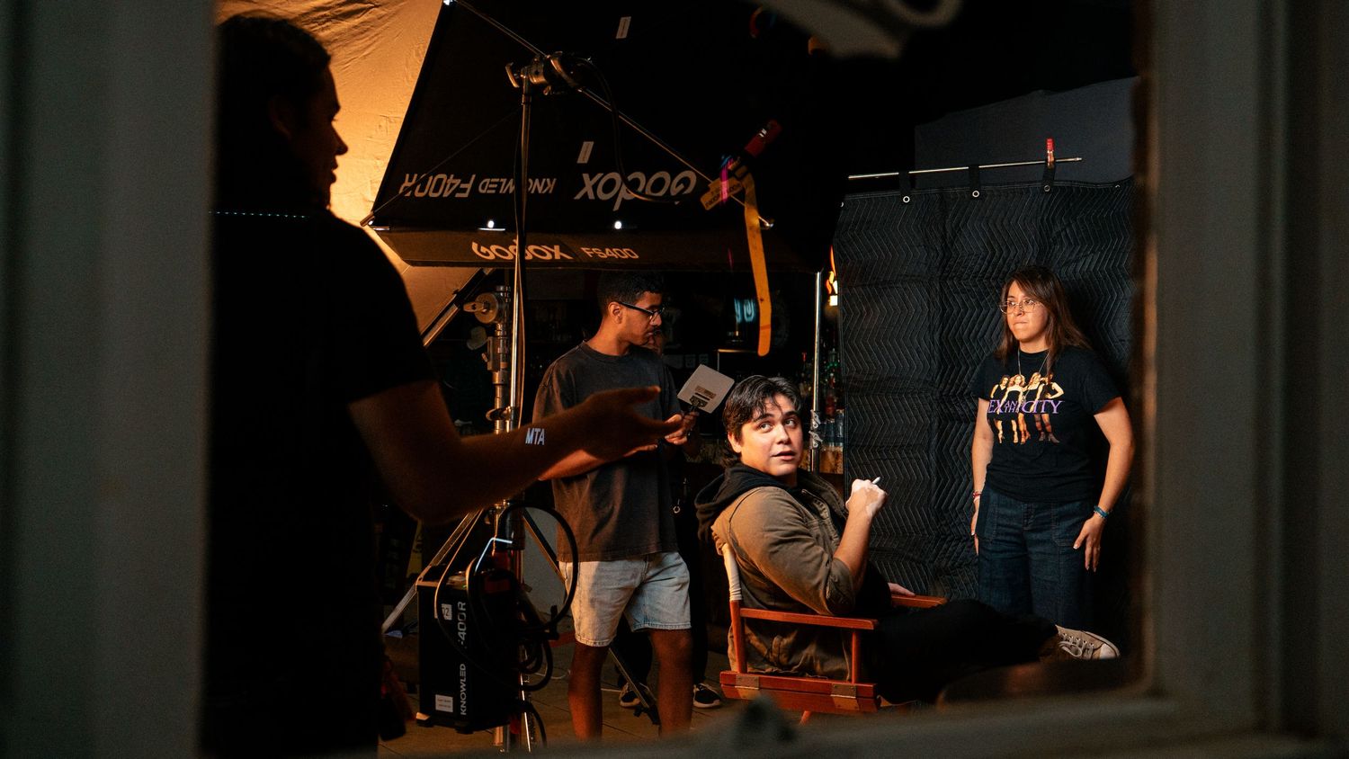 A film crew sets up lighting equipment while directing an actress on an indoor set.