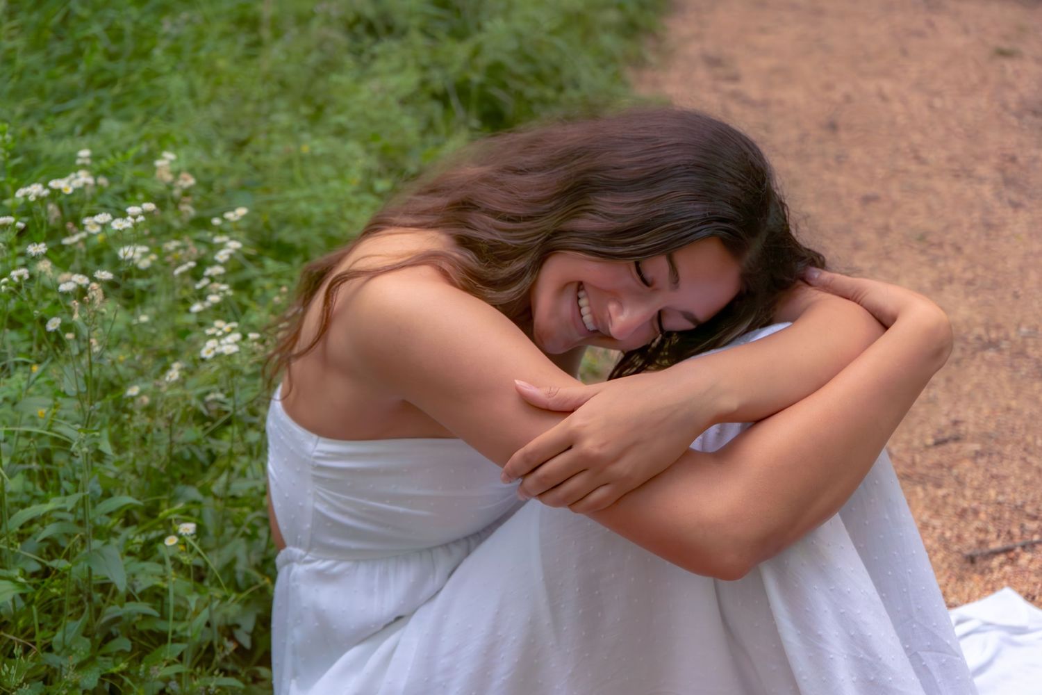 Happy young woman in white dress sitting on path hugging knees among wildflowers.