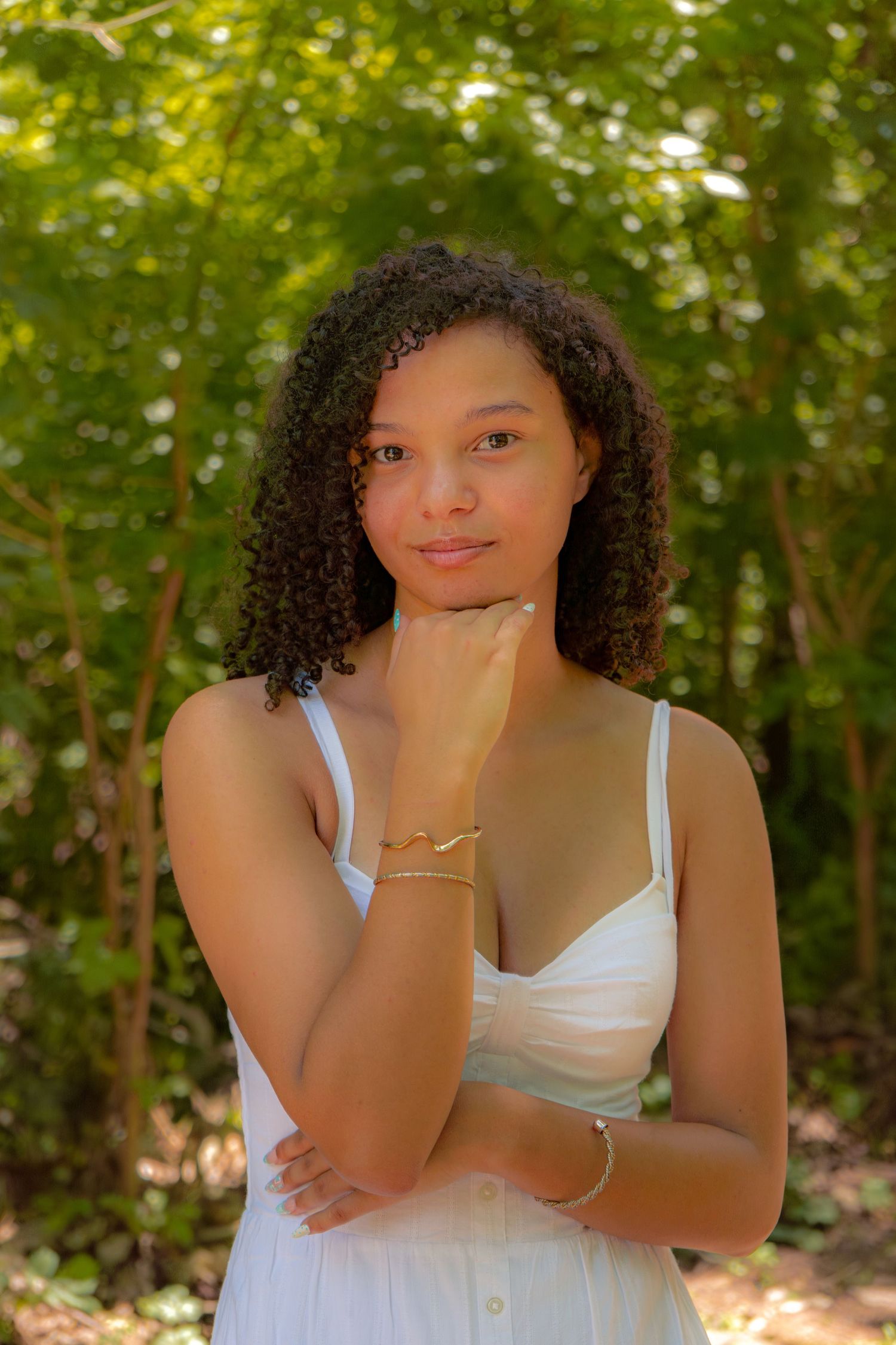 Young woman in white dress poses thoughtfully outdoors, resting chin on hand, wearing gold bracelets.