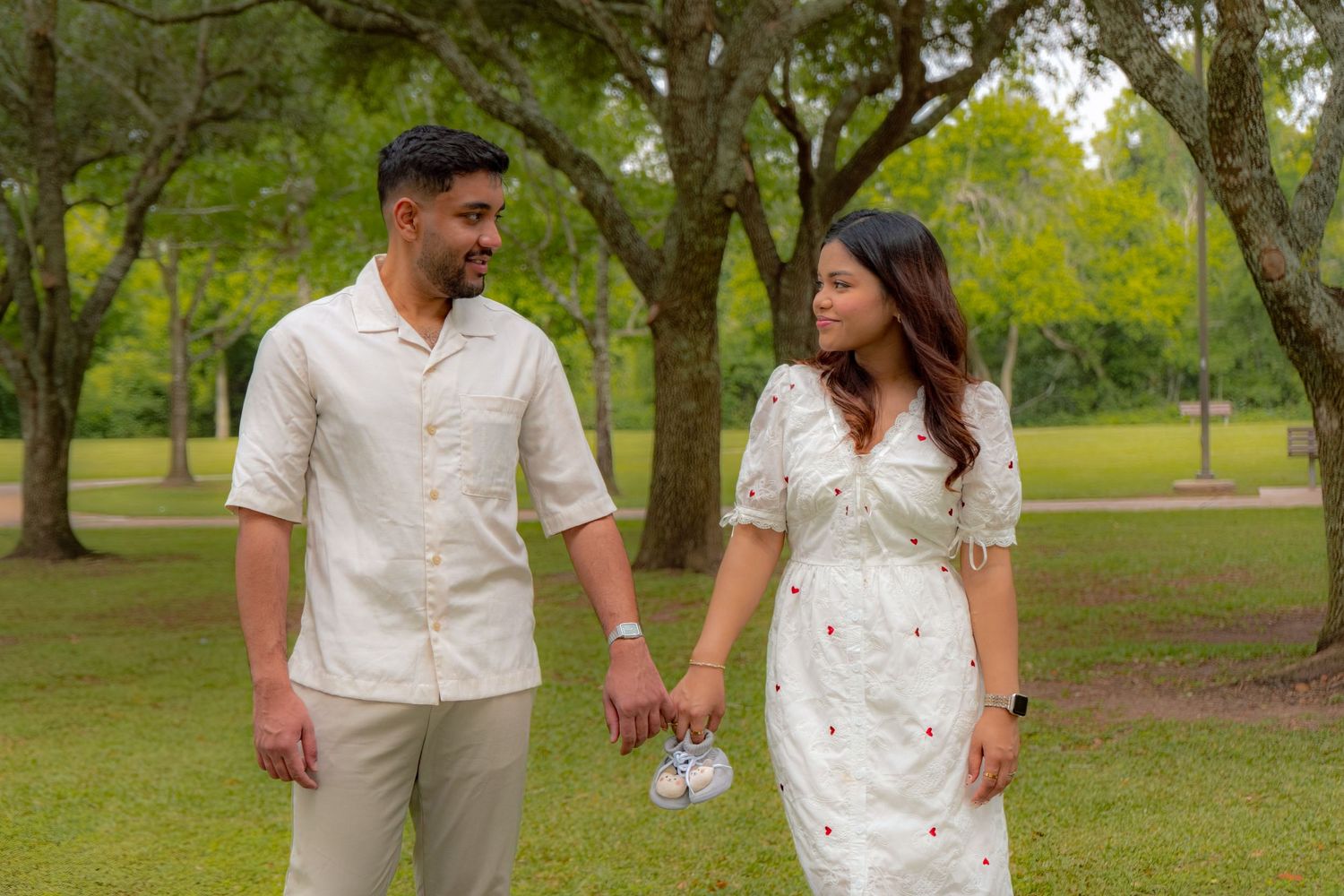 Couple holding hands and smiling at each other while walking in a lush green park surrounded by trees.