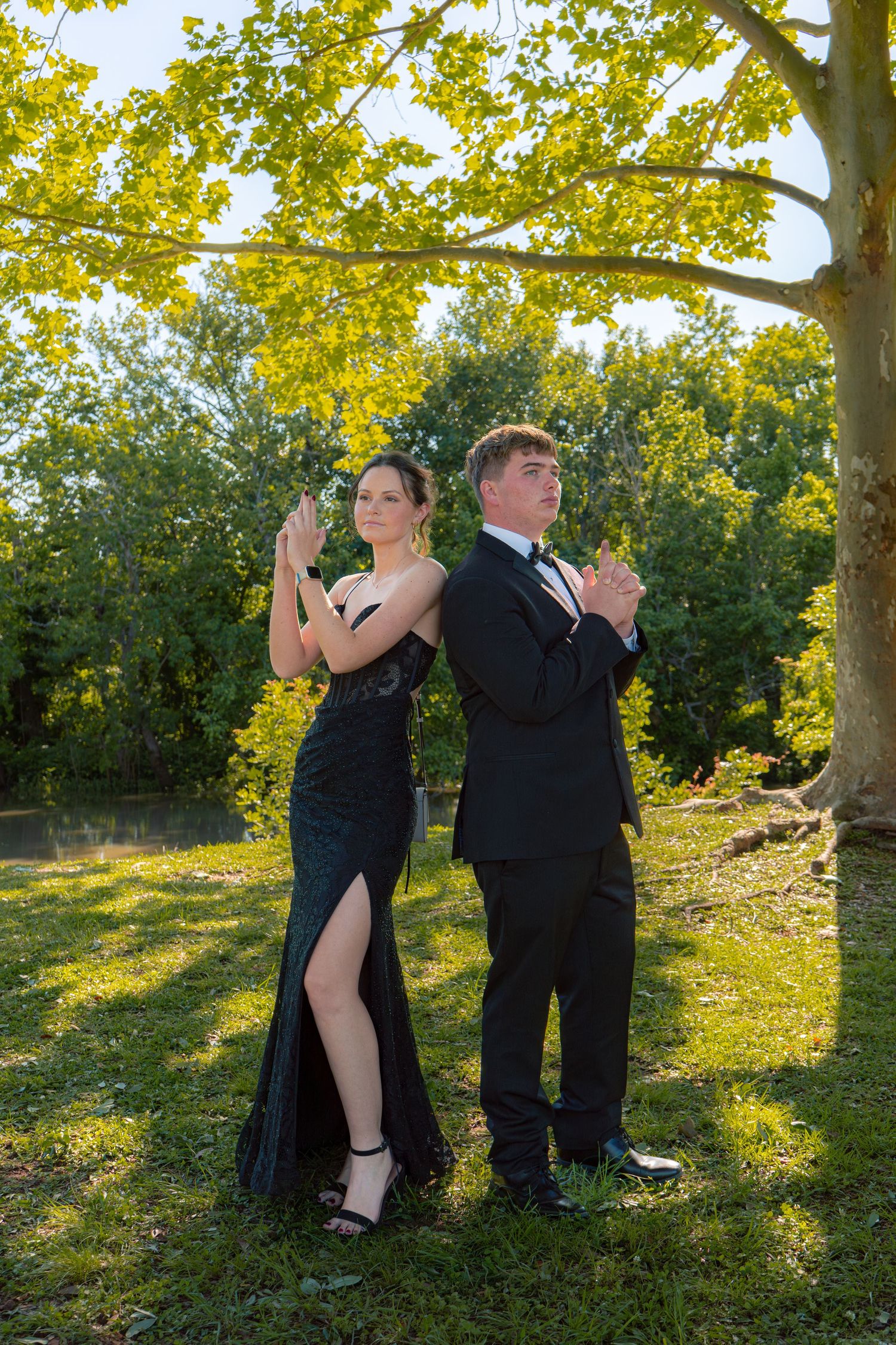 A couple in formal prom attire poses back-to-back under a golden tree in a park.