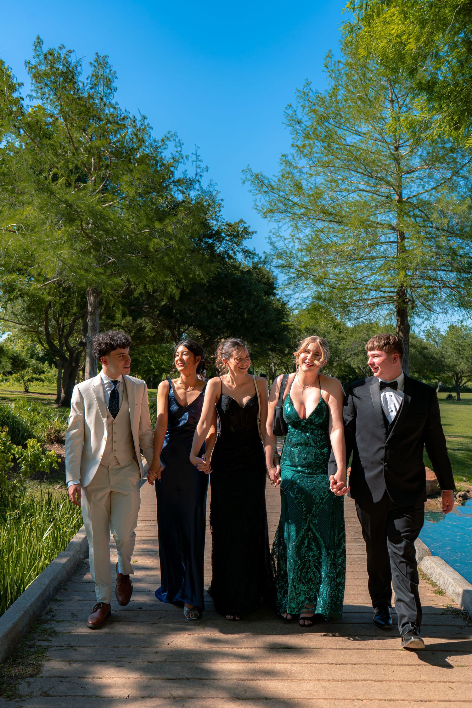 Five teens in formal prom attire walk hand-in-hand along a wooden bridge path surrounded by lush green trees.