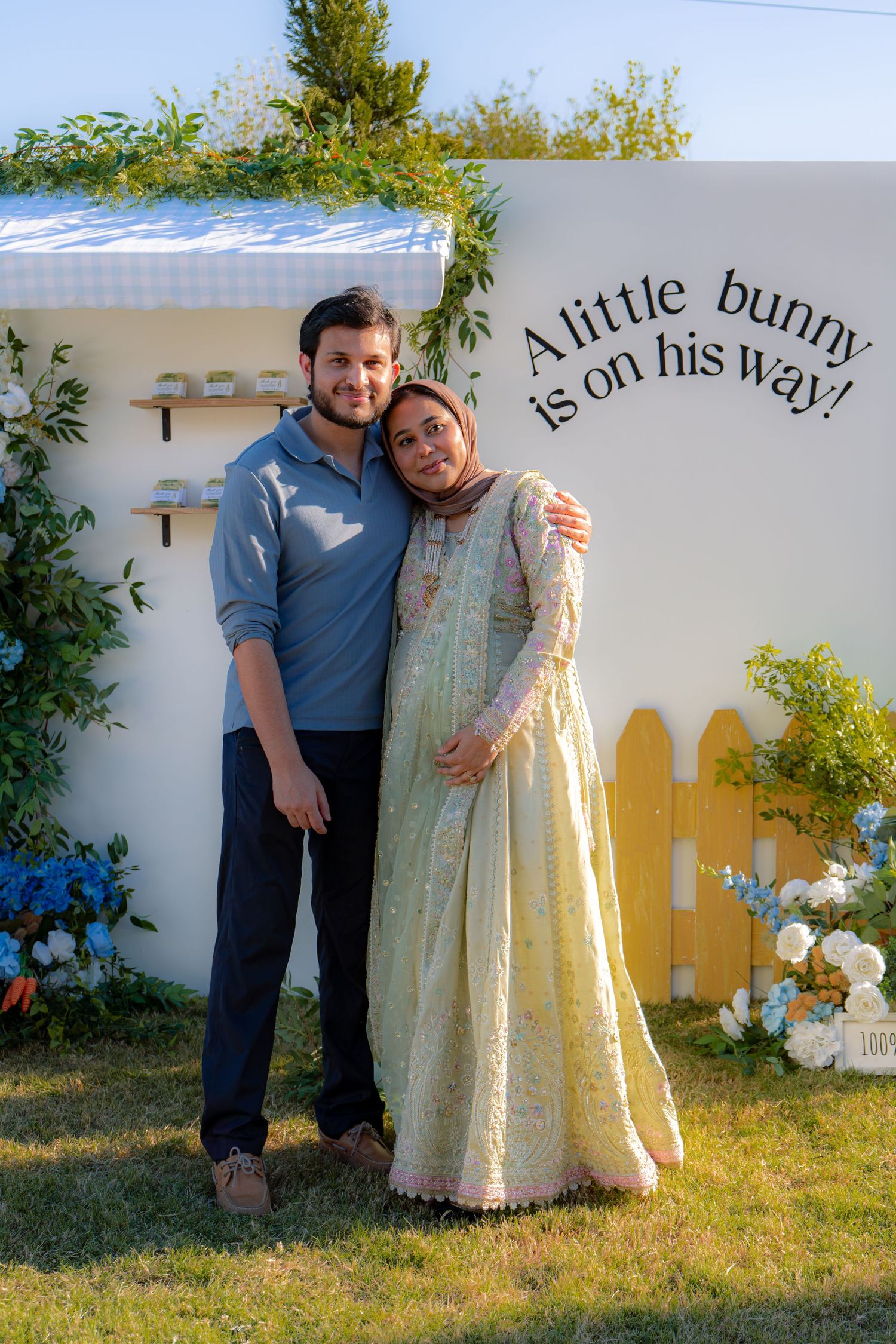 Couple embracing at Peter Rabbit baby shower with white wall sign and yellow fence backdrop.