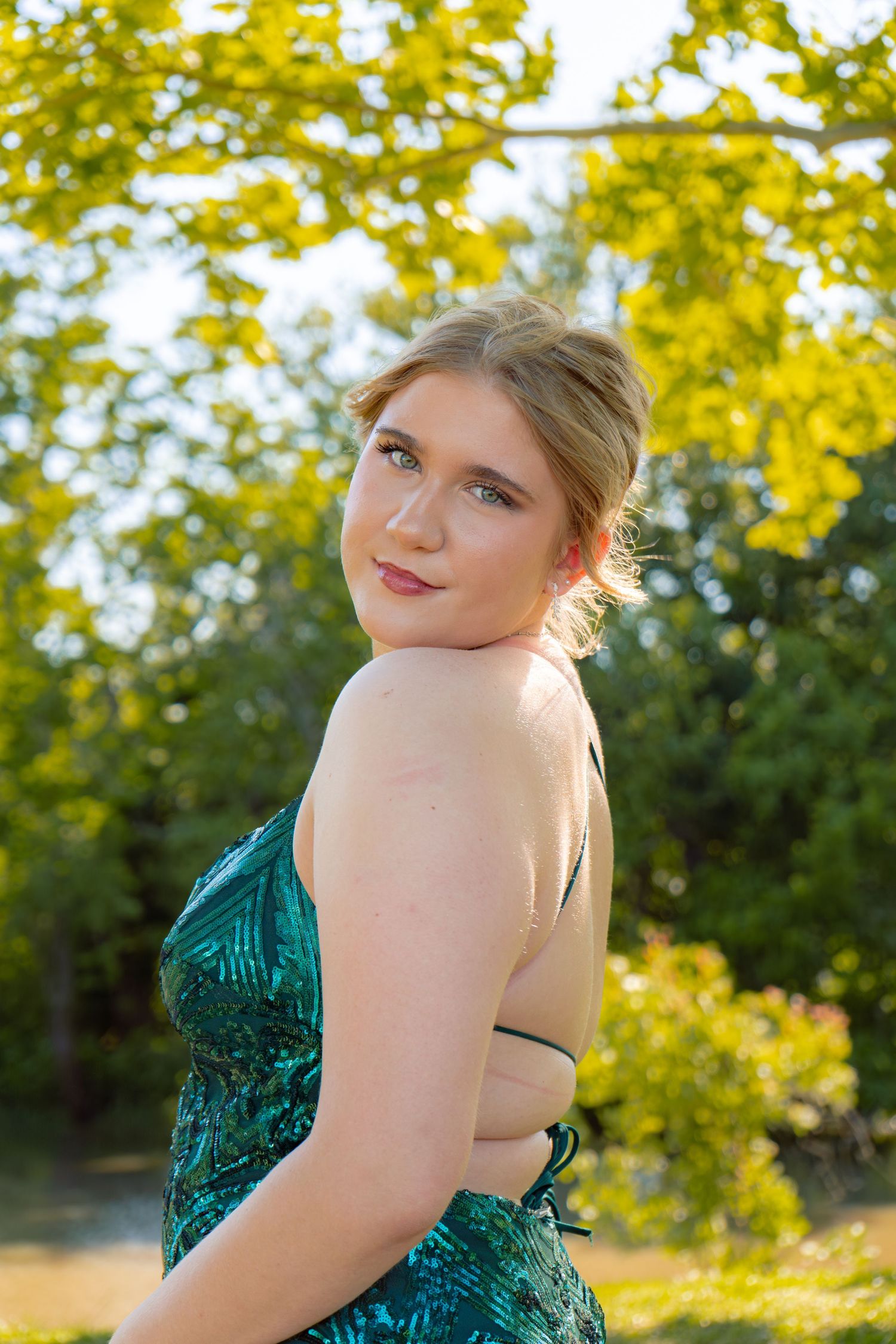 Young woman in a backless green sequin gown poses outdoors with lush yellow-green trees in background.