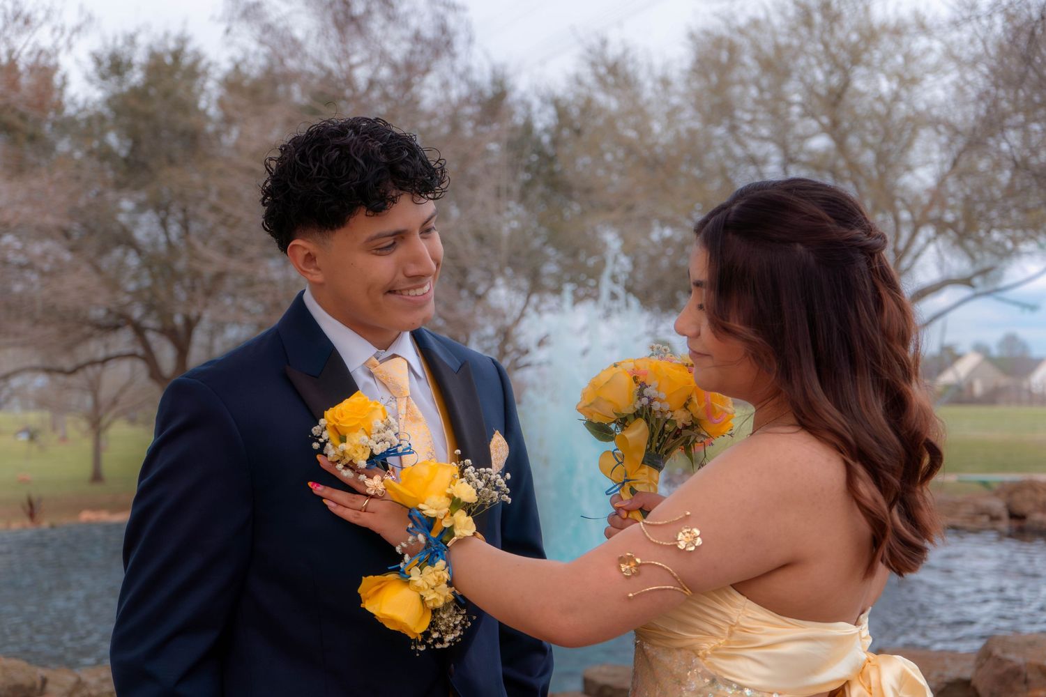 Prom couple sharing a smile while exchanging corsage and bouquet of yellow roses outdoors.