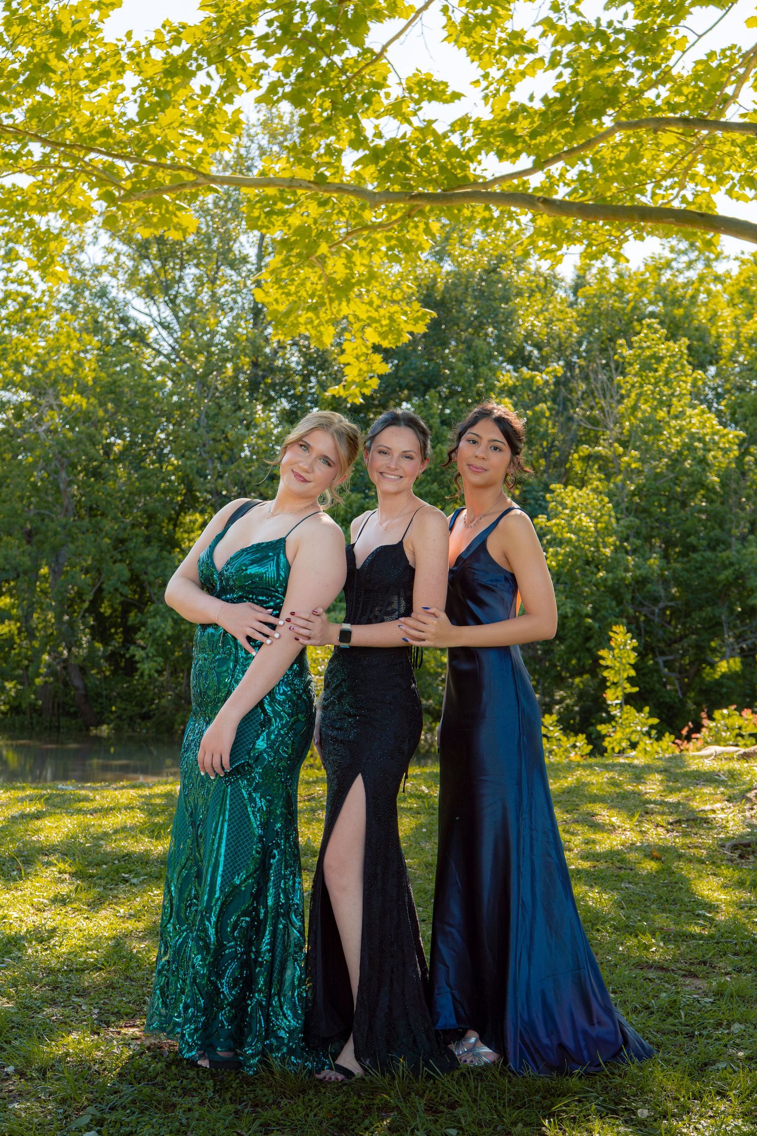 Three young women in elegant prom gowns of green, black, and navy pose together outdoors under yellow-green tree branches.