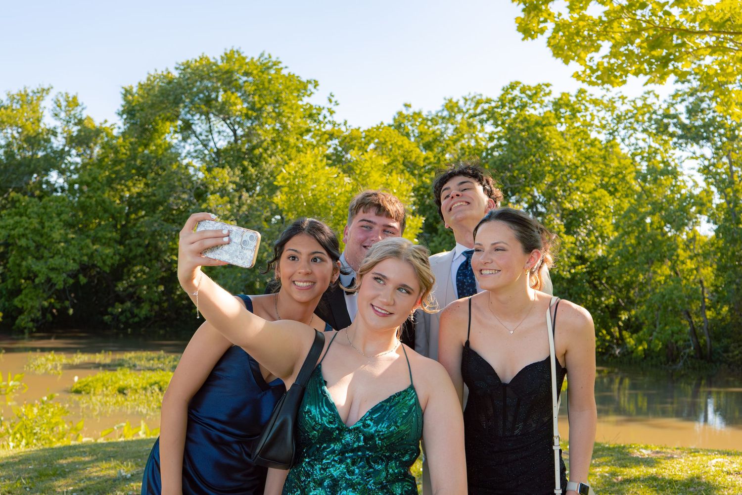Group of five prom-dressed teens take selfie outdoors near a pond.