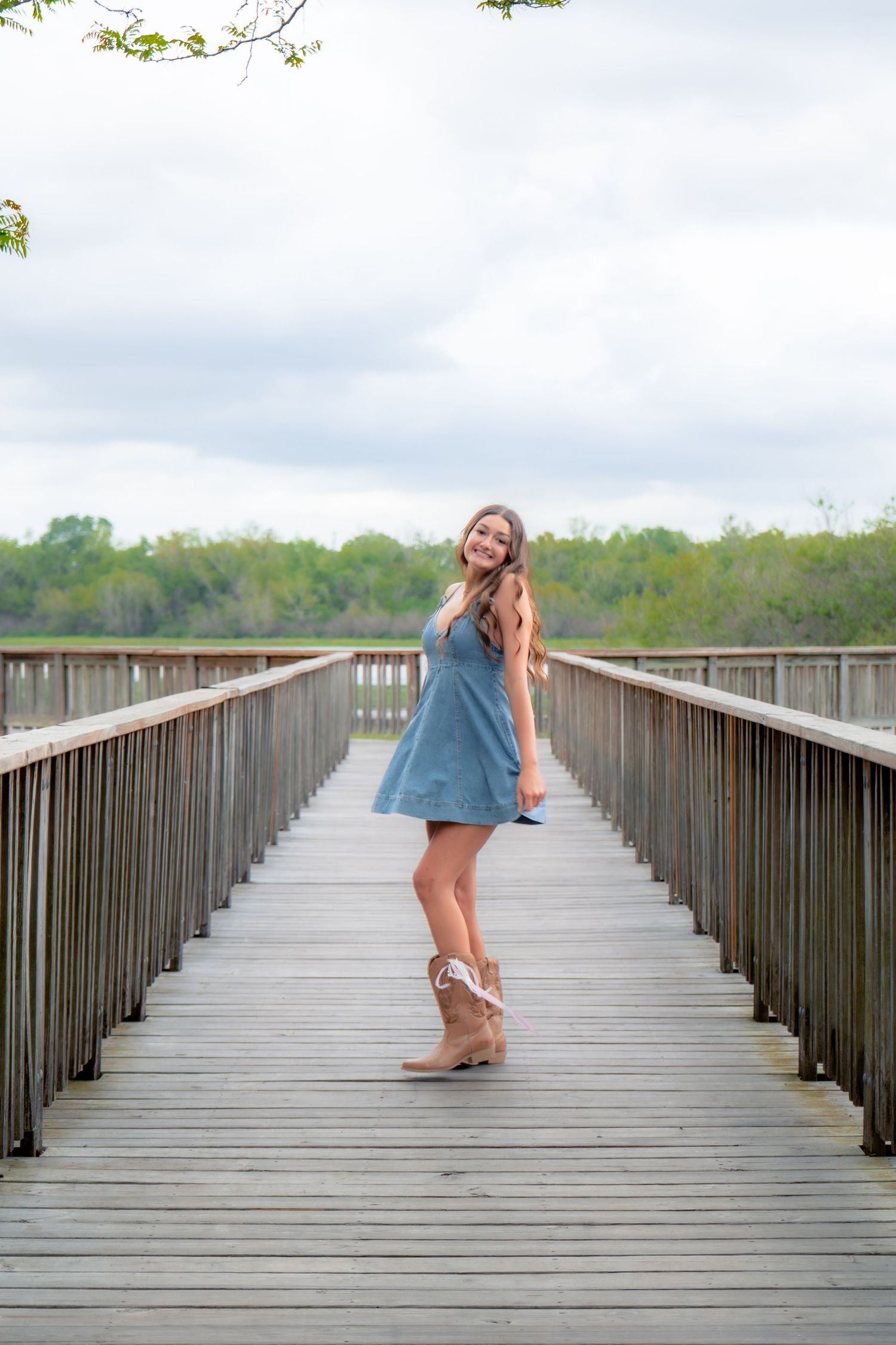 Smiling young woman in blue dress and tan boots posing on a wooden bridge surrounded by lush greenery.