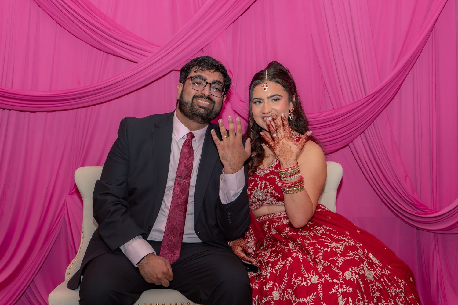 Indian couple in traditional wedding attire showing henna hands and rings against pink backdrop.