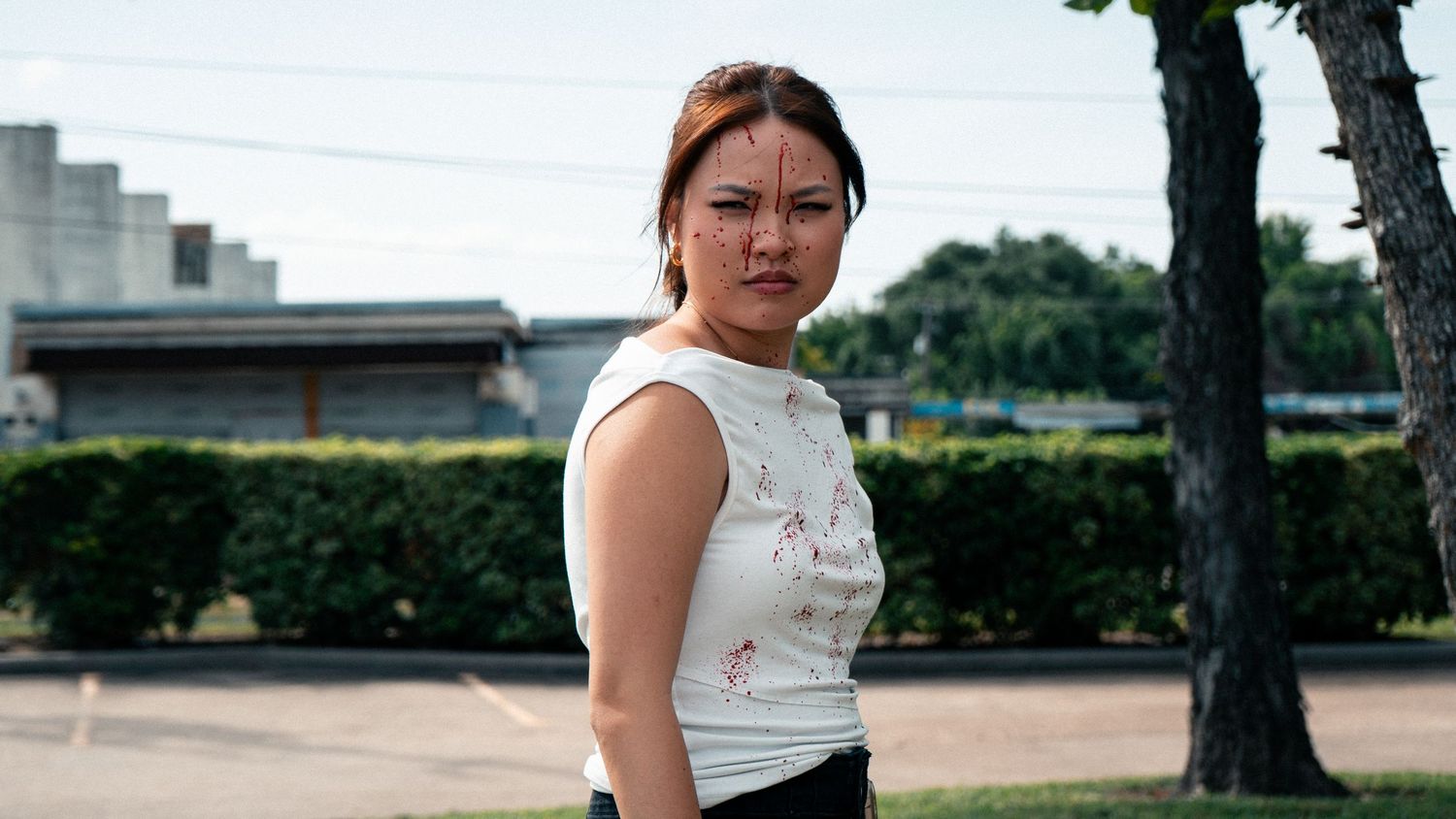 Young Asian woman with face markings stands outdoors in a white top, looking serious.