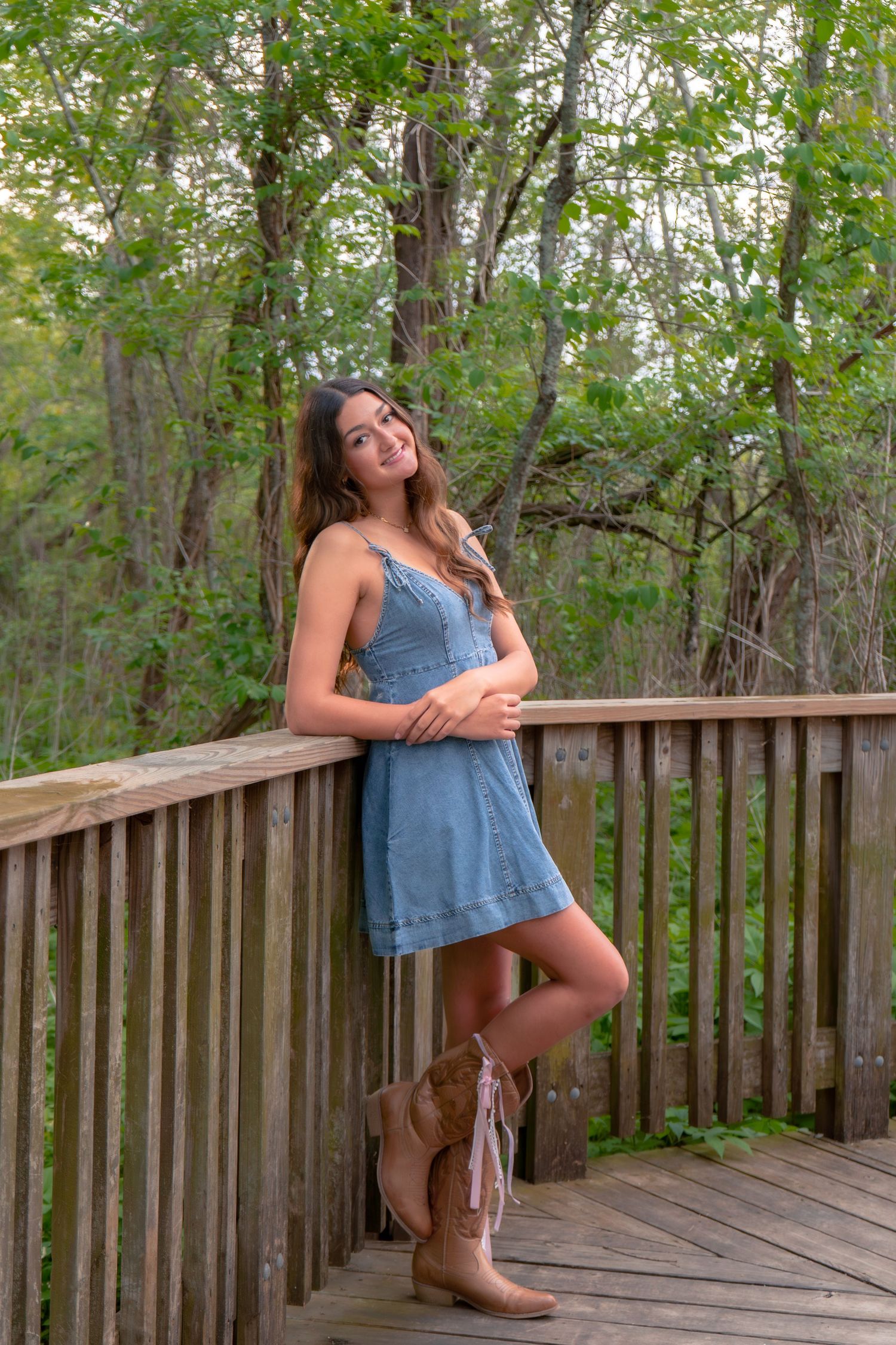 Young woman in denim dress and cowboy boots leaning on wooden deck railing surrounded by lush green trees.
