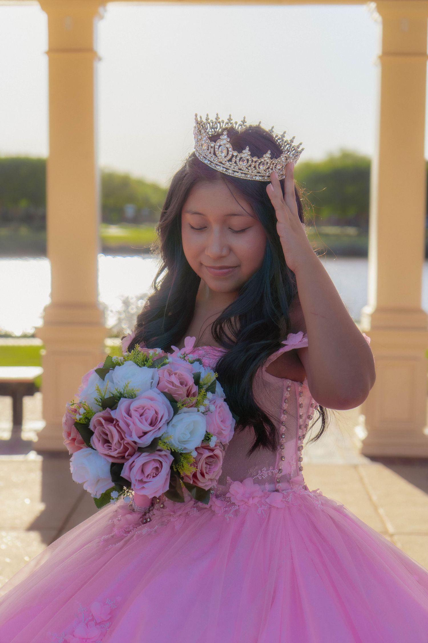 Young Latina in pink quinceañera gown holds floral bouquet while adjusting tiara crown outdoors near water.