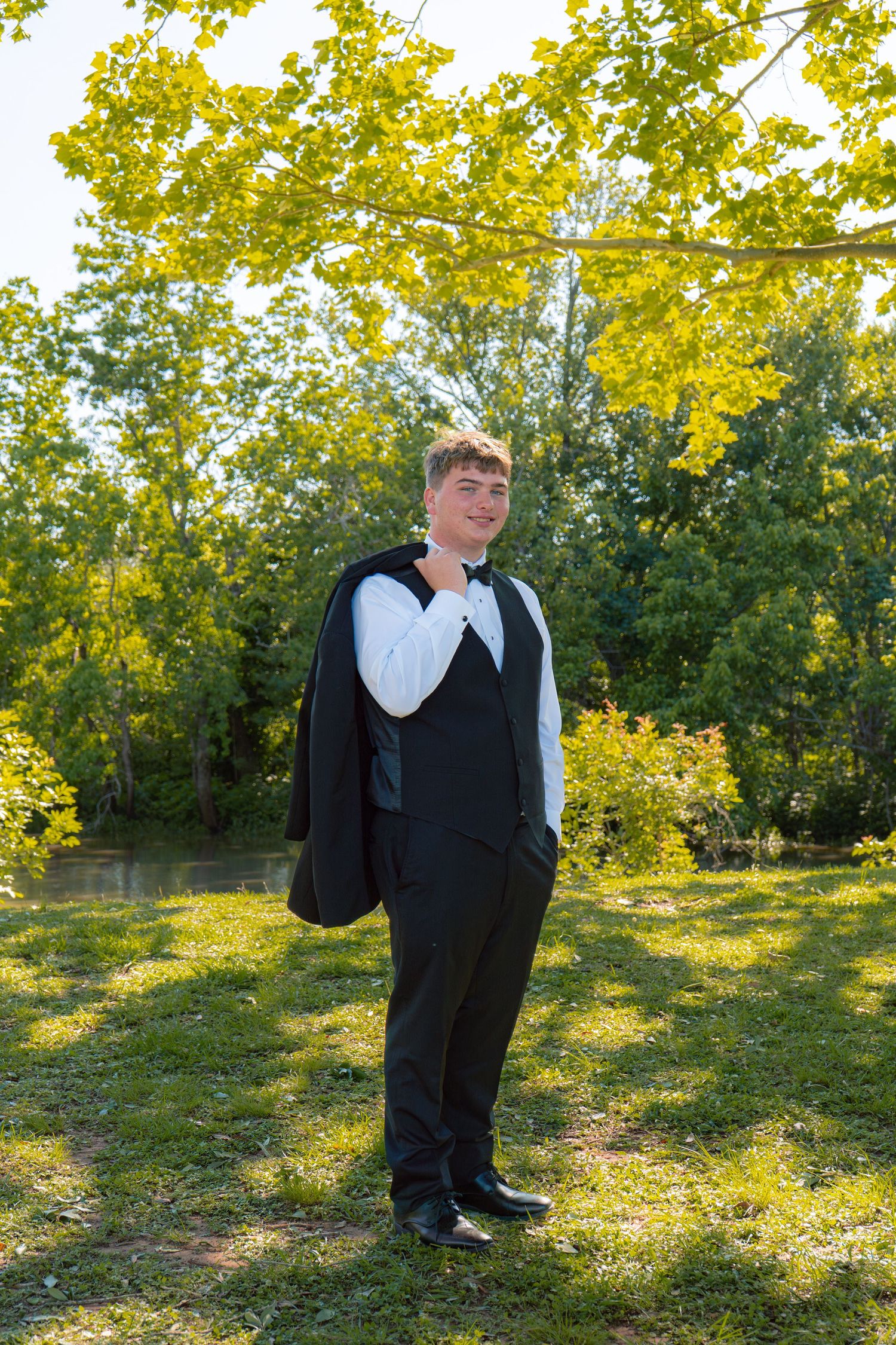 Young man in black tuxedo vest carries jacket over shoulder while posing outdoors in sunny garden.
