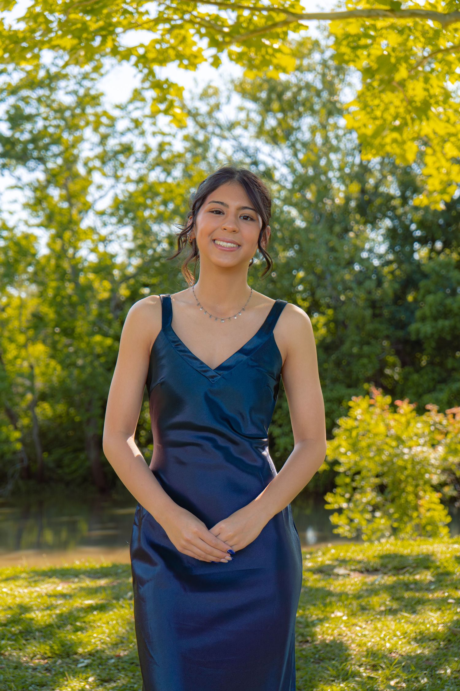 Young woman in navy satin gown smiling outdoors in lush green park setting.