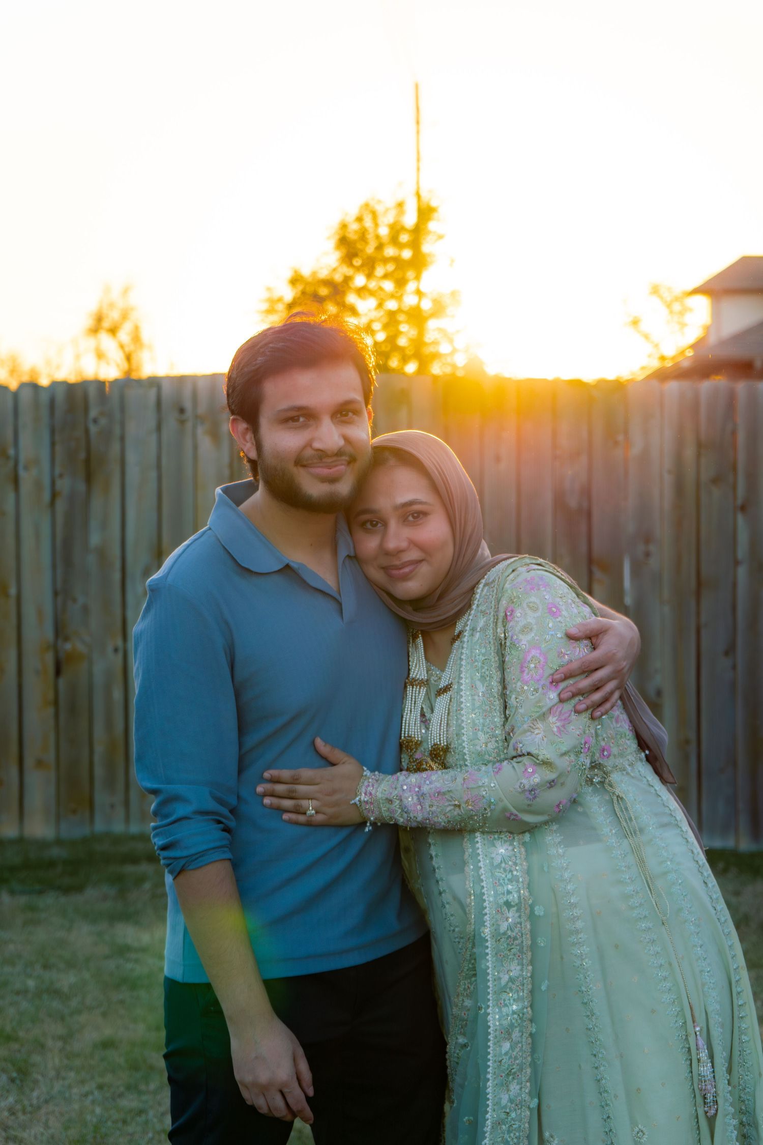 Couple embracing outdoors at golden sunset, woman wearing hijab and floral dress, man in blue polo shirt, wooden fence background.