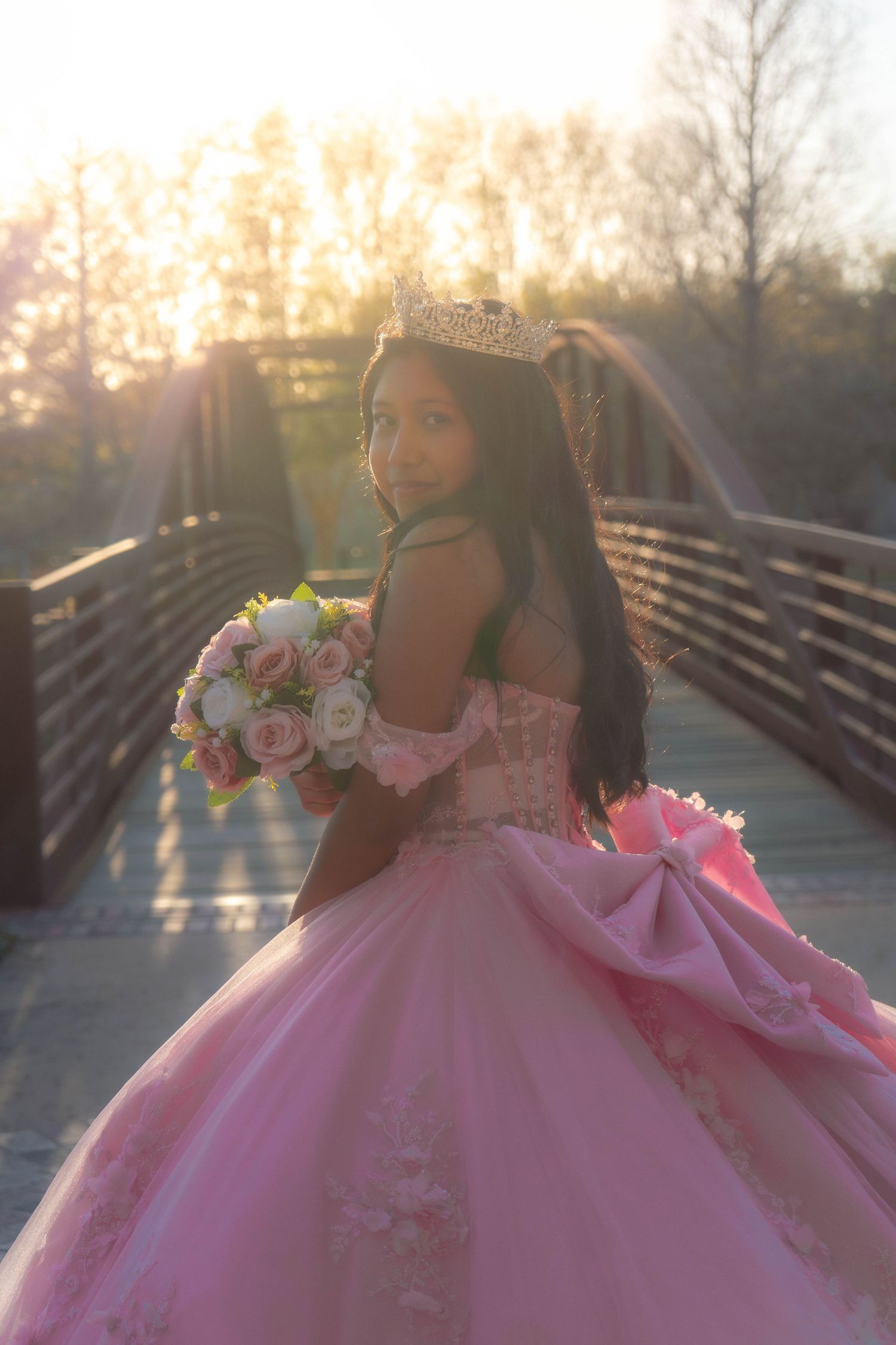 Teen girl in pink ball gown quinceañera dress holding bouquet on bridge at golden hour sunset.