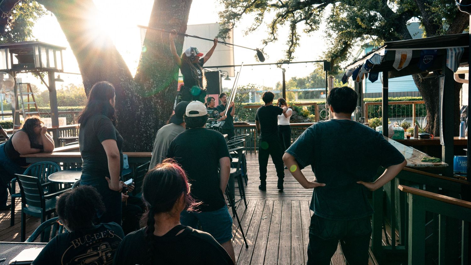 A film crew sets up an outdoor shot on a wooden deck surrounded by trees.