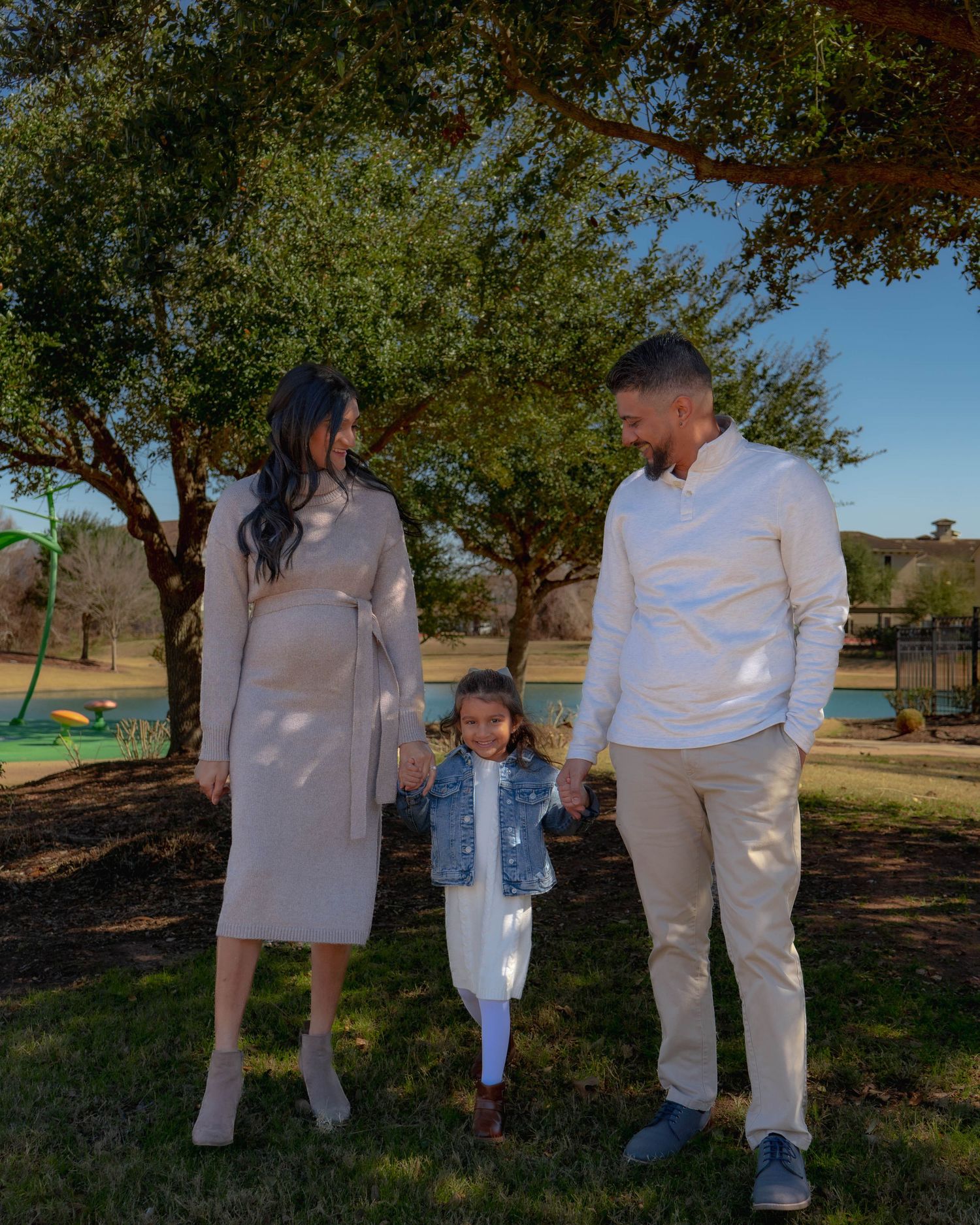 Happy family maternity portrait with parents and toddler holding hands in park.