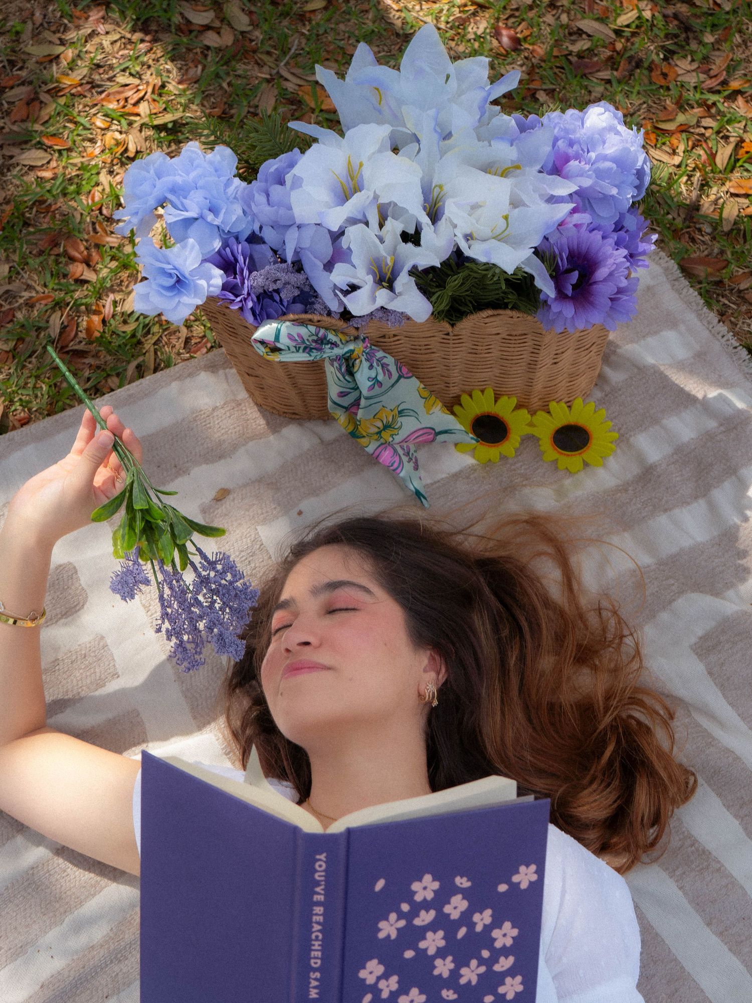 Woman lying outdoors reading floral purple book, holding lavender stems beside sunflowers and blue flower basket.