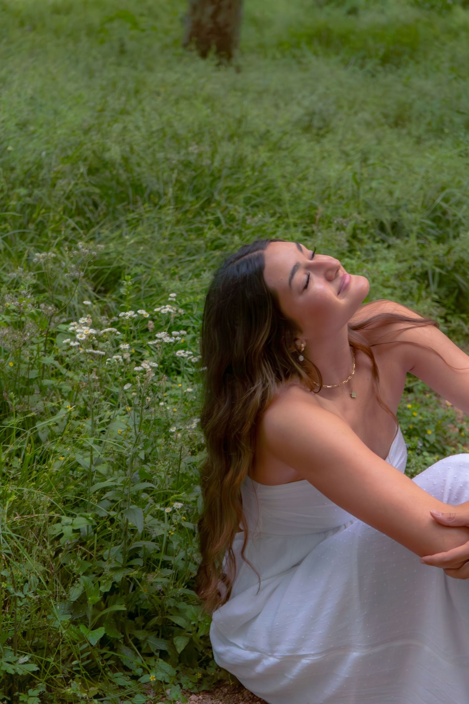 Young woman in white dress sitting in green meadow with eyes closed, head tilted back.
