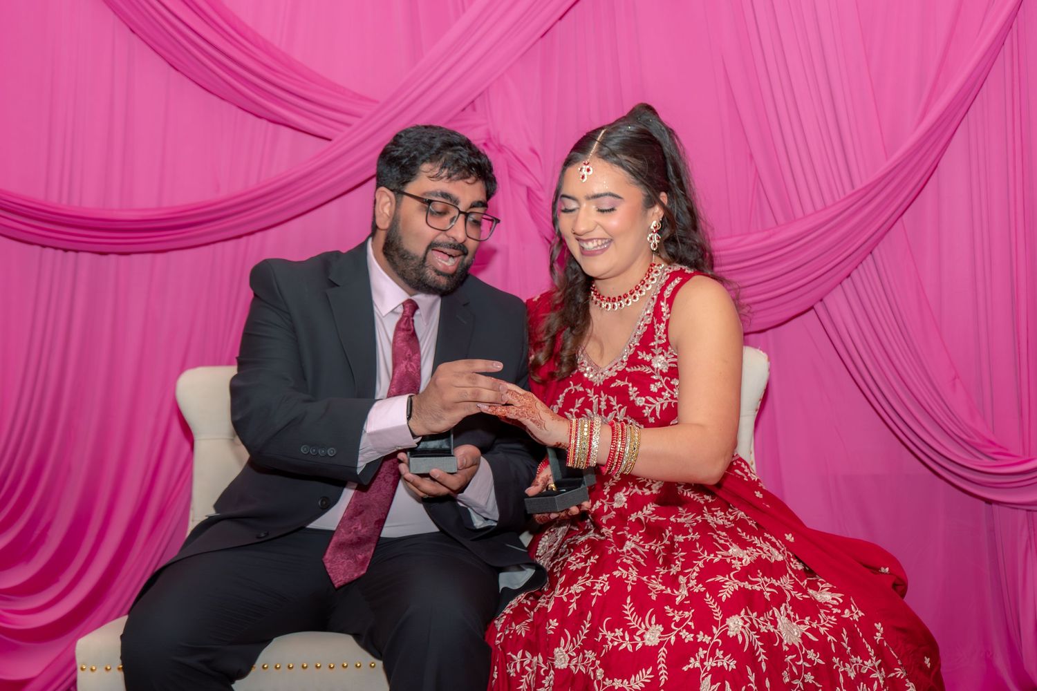 South Asian couple seated on sofa examining ring box, bride in red embroidered lehenga against pink draped backdrop.