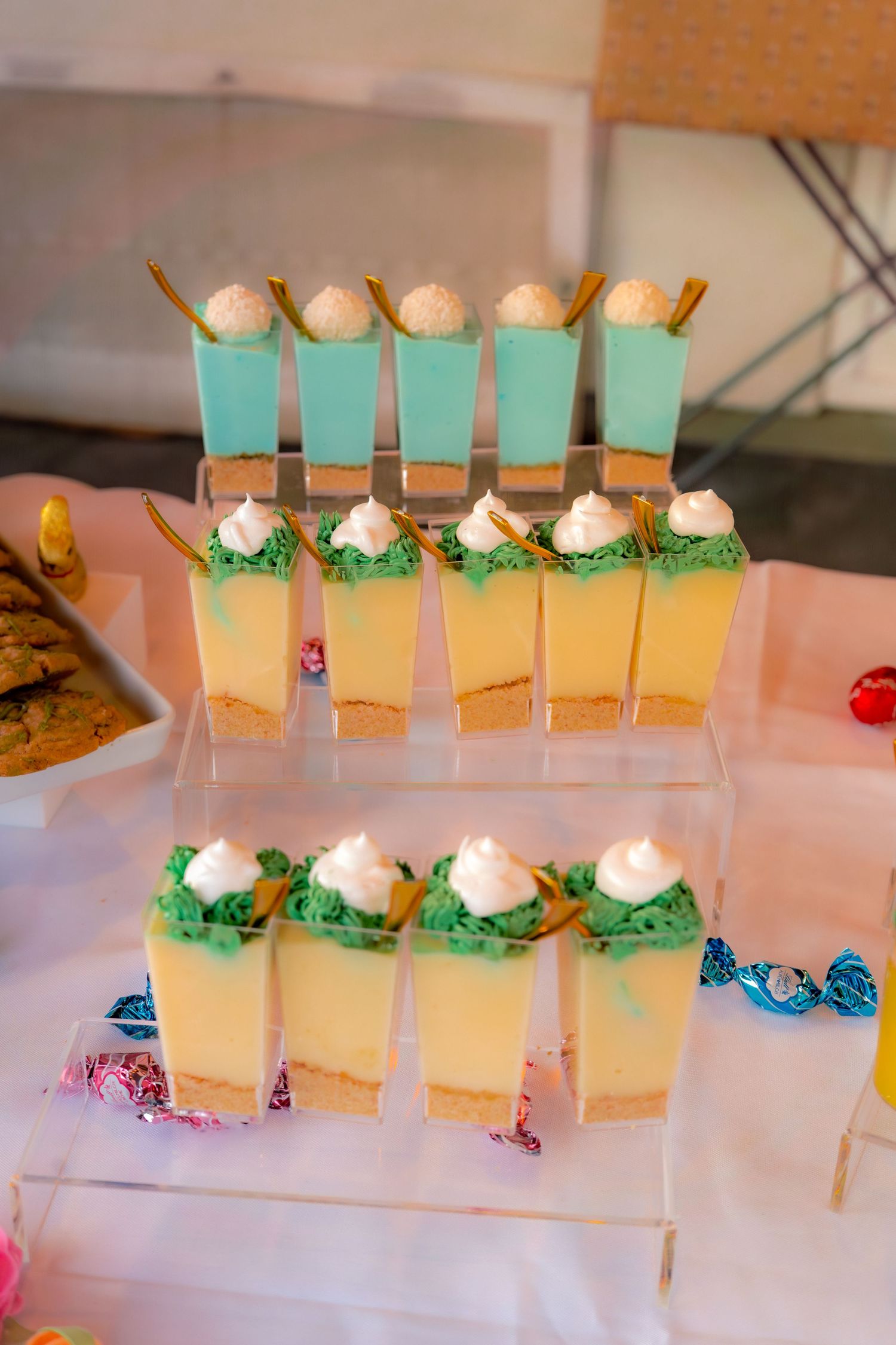 Layered yellow and green mini dessert cups with white cream topping displayed on a clear acrylic tiered stand.