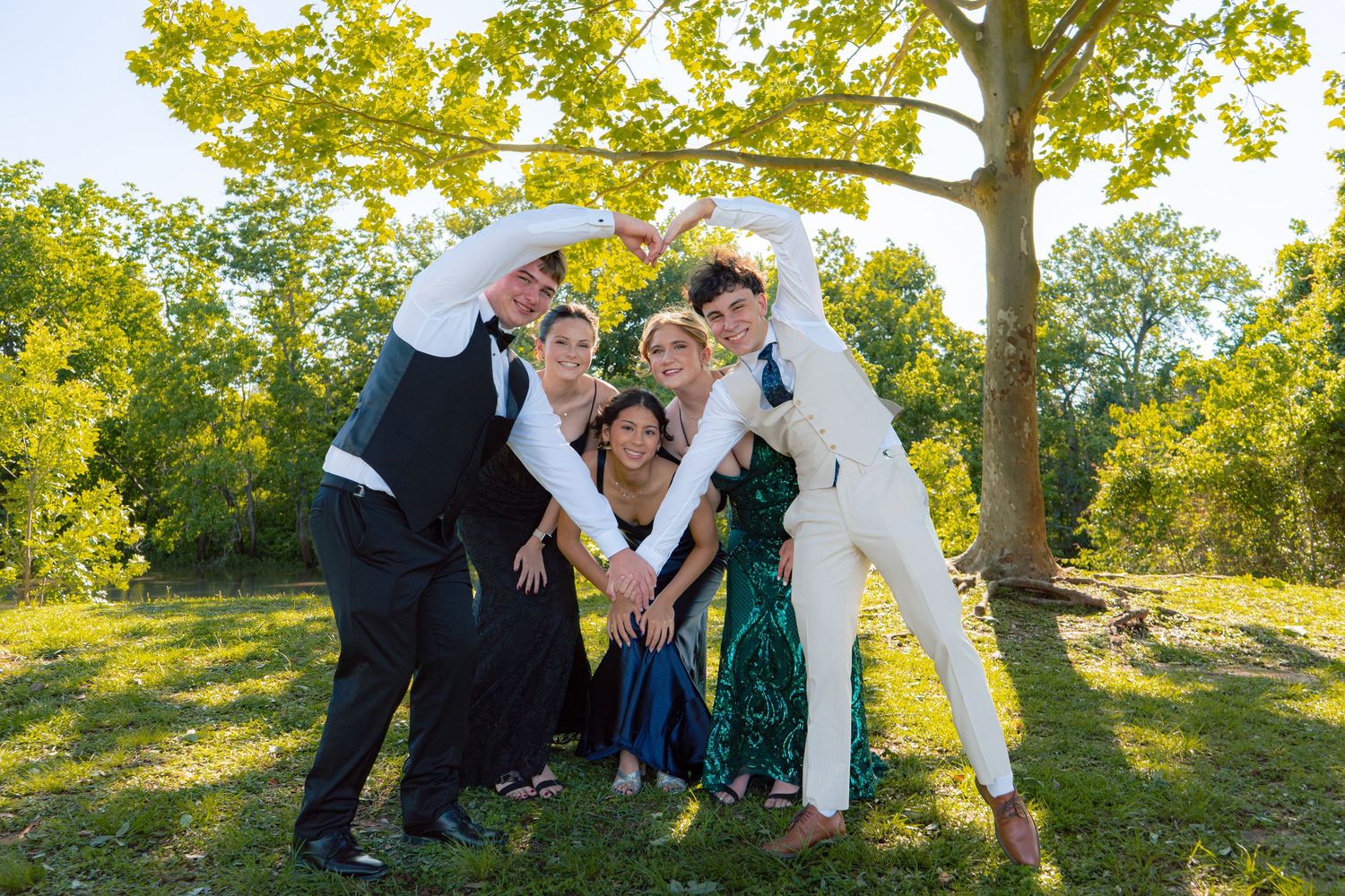 A group of six formally dressed teens pose playfully under a tree in a sunny park.
