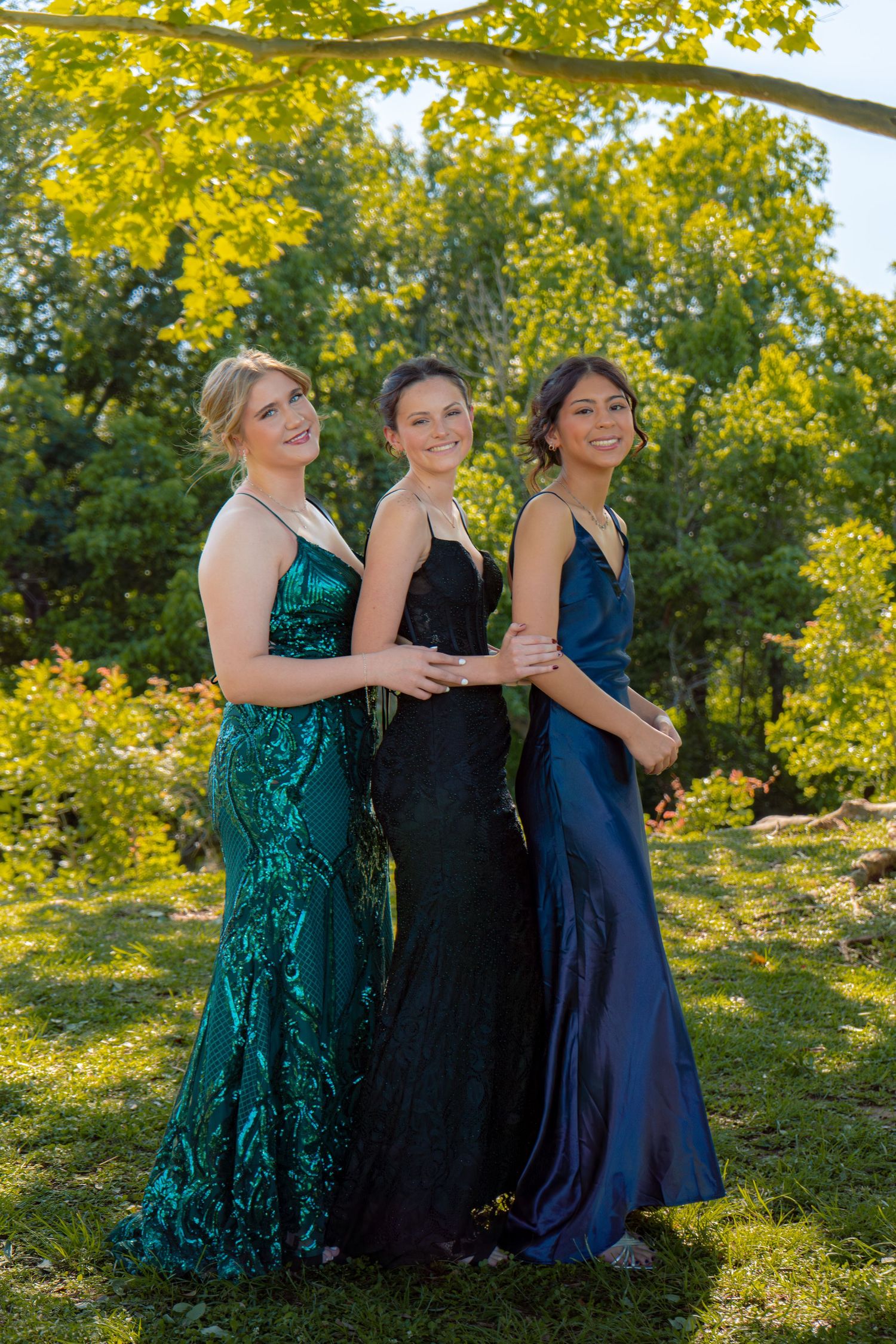 Three young women in elegant prom gowns of green, black, and navy blue pose outdoors in a lush garden setting.