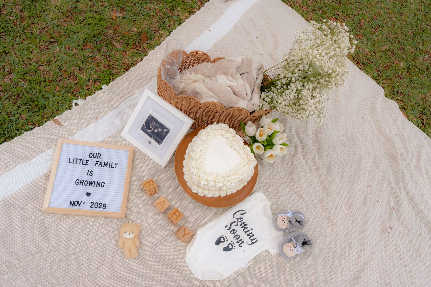 Flat lay pregnancy announcement with Coming Soon onesie, ultrasound photo, letter board, and flowers on beige blanket.