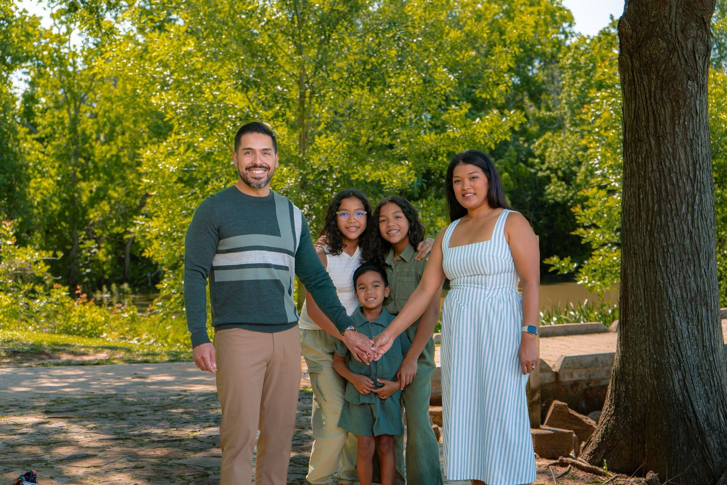 Family of five smiling outdoors in a lush green park setting on a sunny day.
