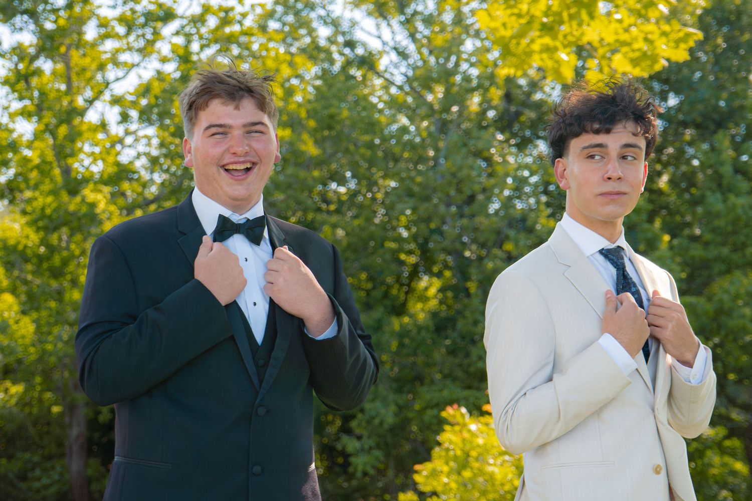 Two young men in tuxedos adjusting their ties outdoors with green trees in the background.