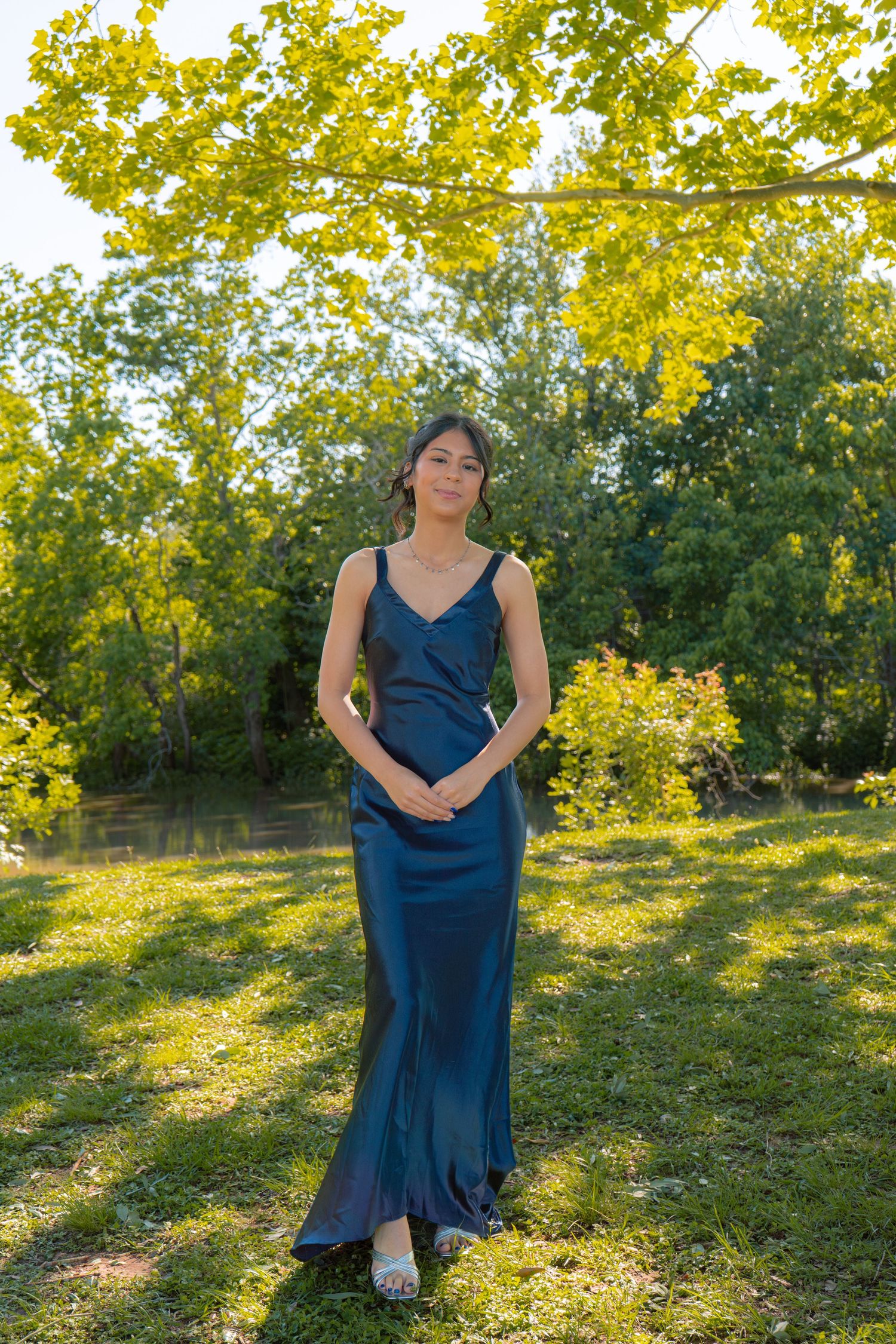 Elegant woman in full-length navy satin gown standing on grass beneath yellow-green trees.