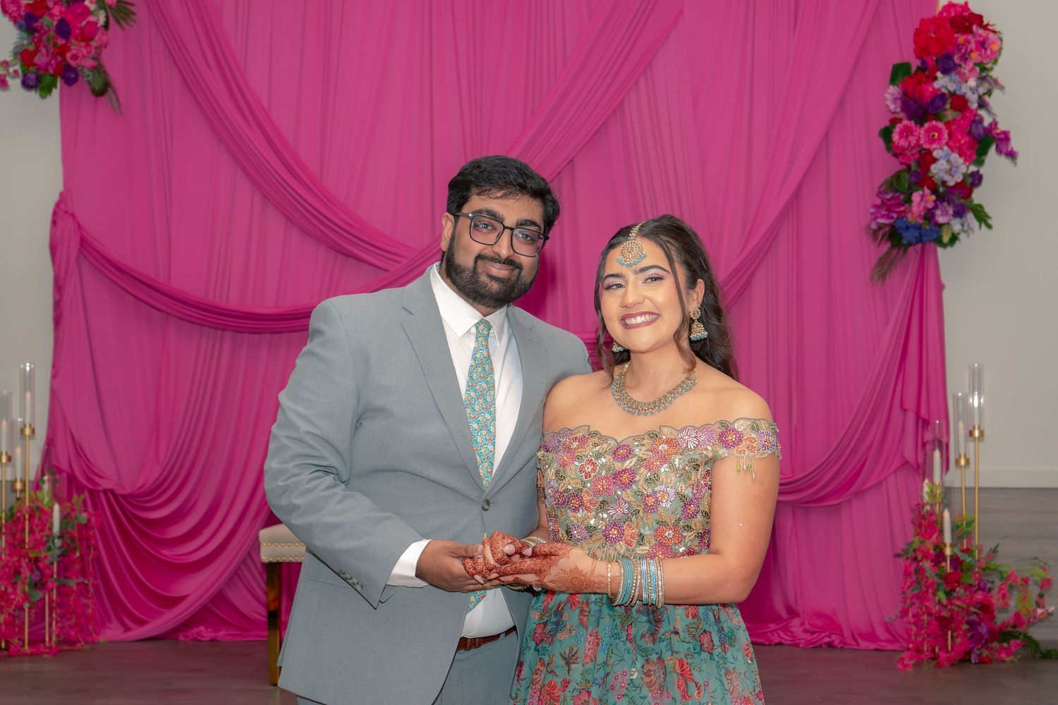 Couple smiling at engagement ceremony with pink floral backdrop, man in gray suit, woman in colorful off-shoulder dress.