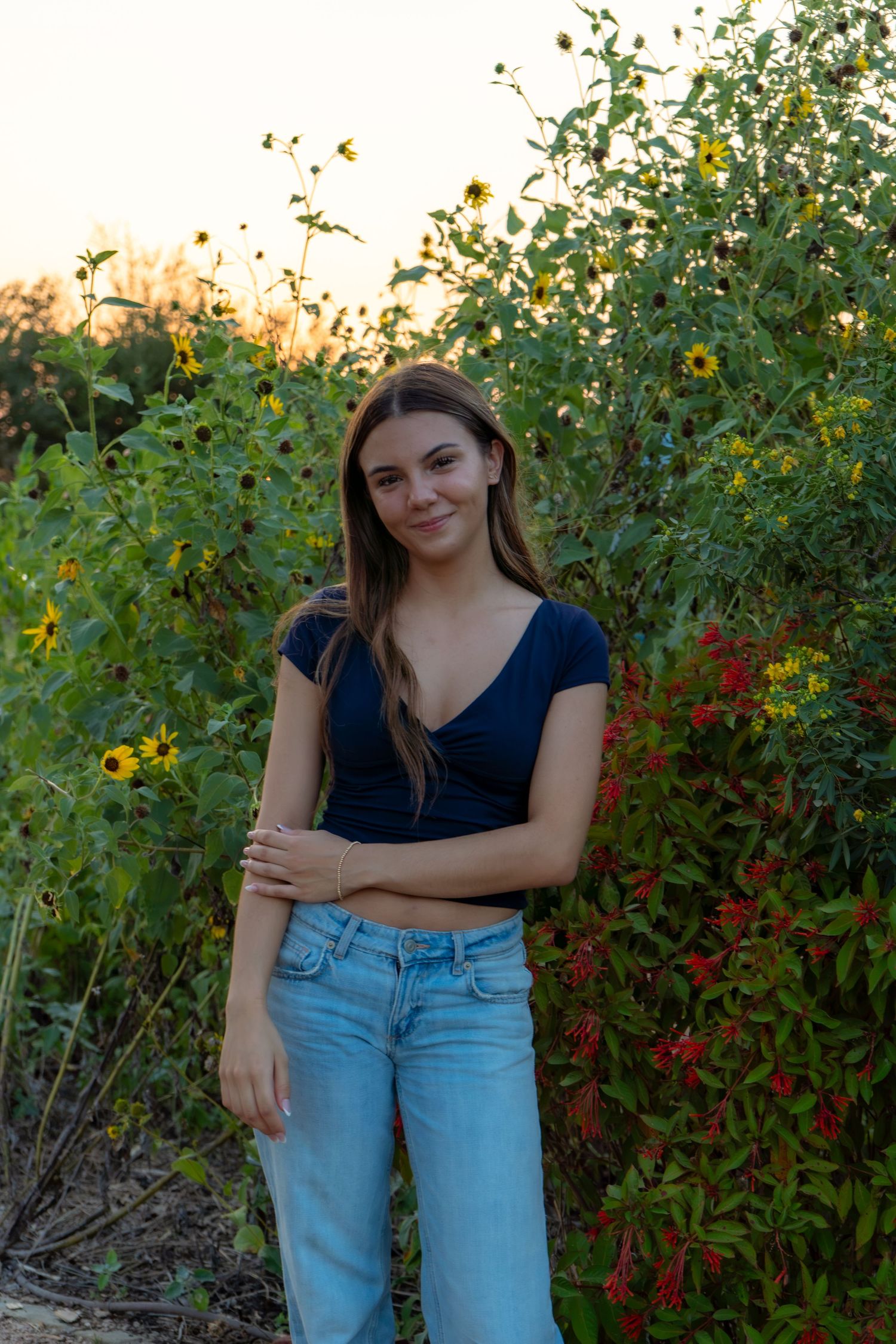Young woman standing among wildflowers and red blooms at sunset, wearing blue crop top and jeans, smiling softly.