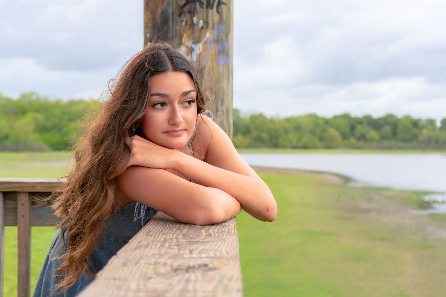 Young woman with long brown hair leaning on wooden railing outdoors near a scenic waterway.