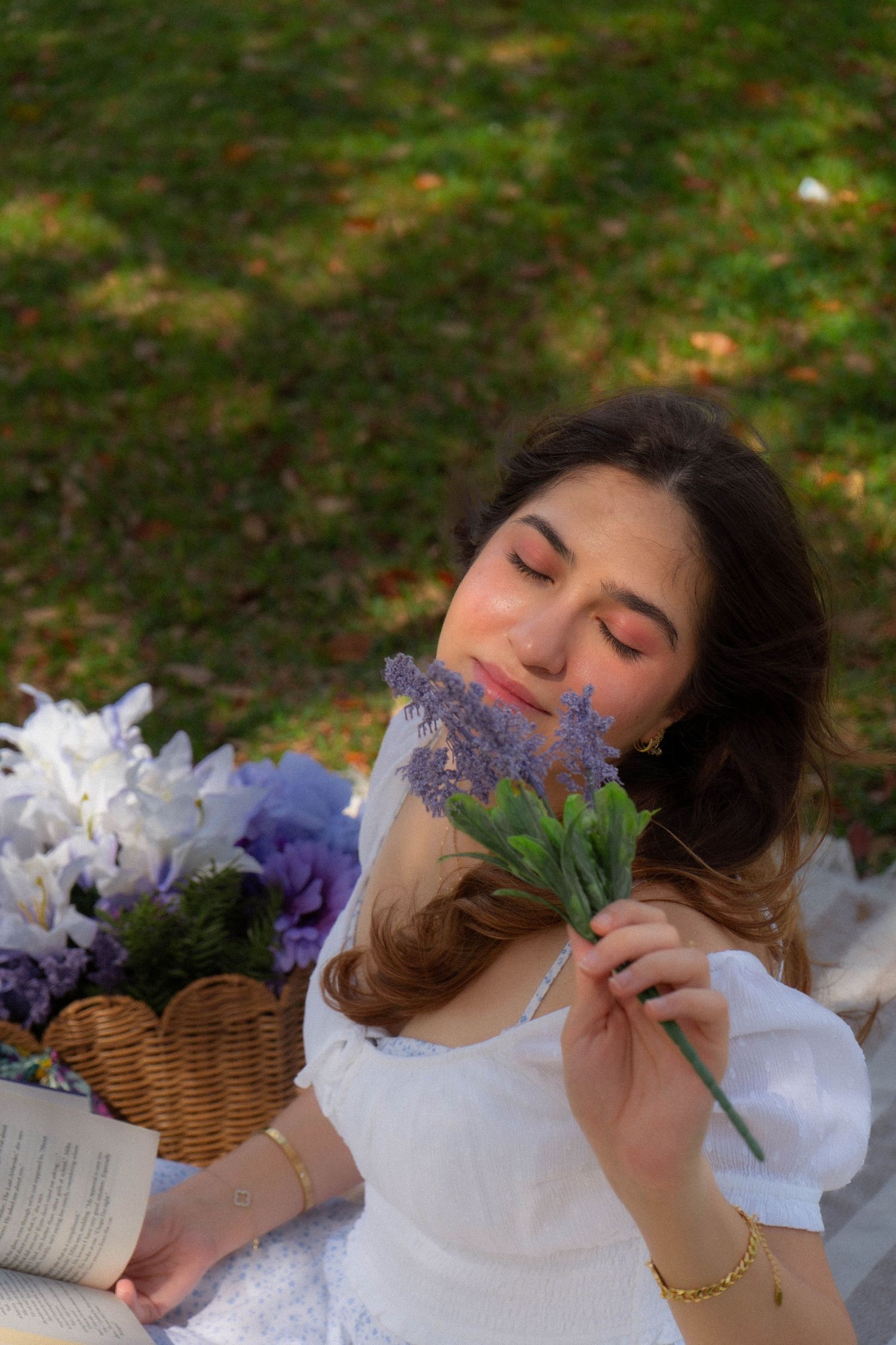 Young woman in white dress smelling lavender flowers with eyes closed near floral basket on grass.