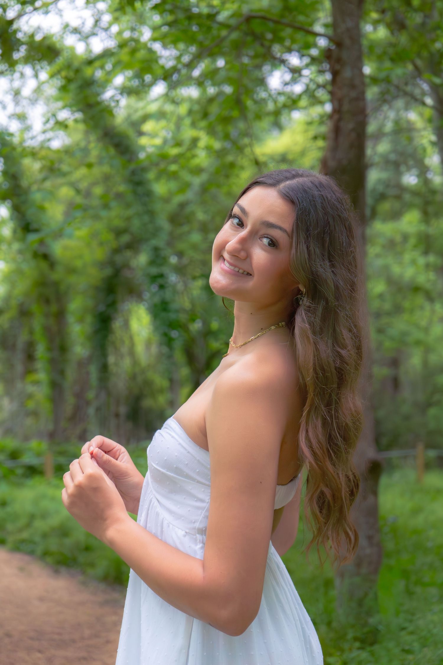 Smiling young woman with long wavy hair wearing a white strapless dress on a forest path.